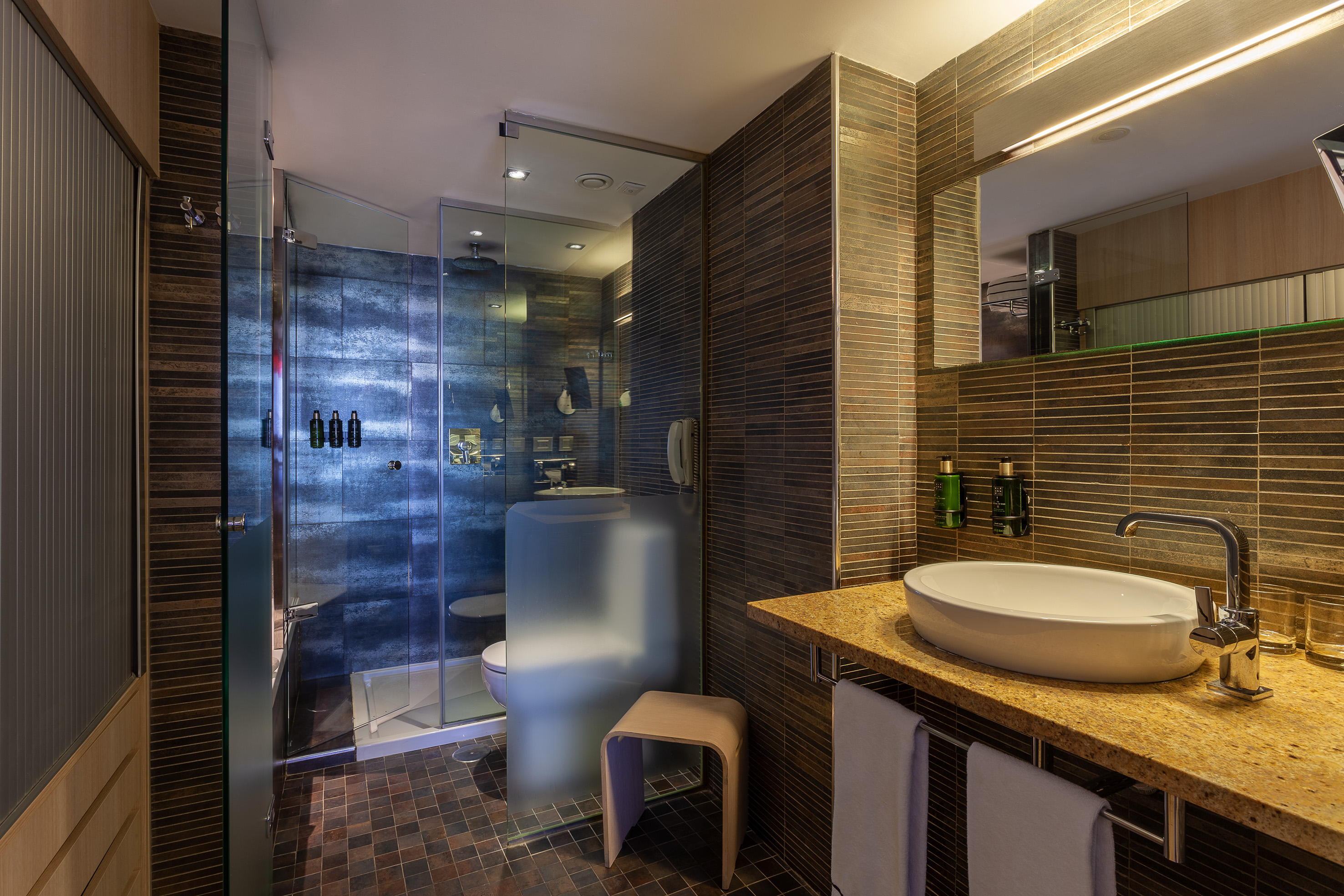 Modern hotel bathroom with glass shower, vessel sink vanity, and dark tile.