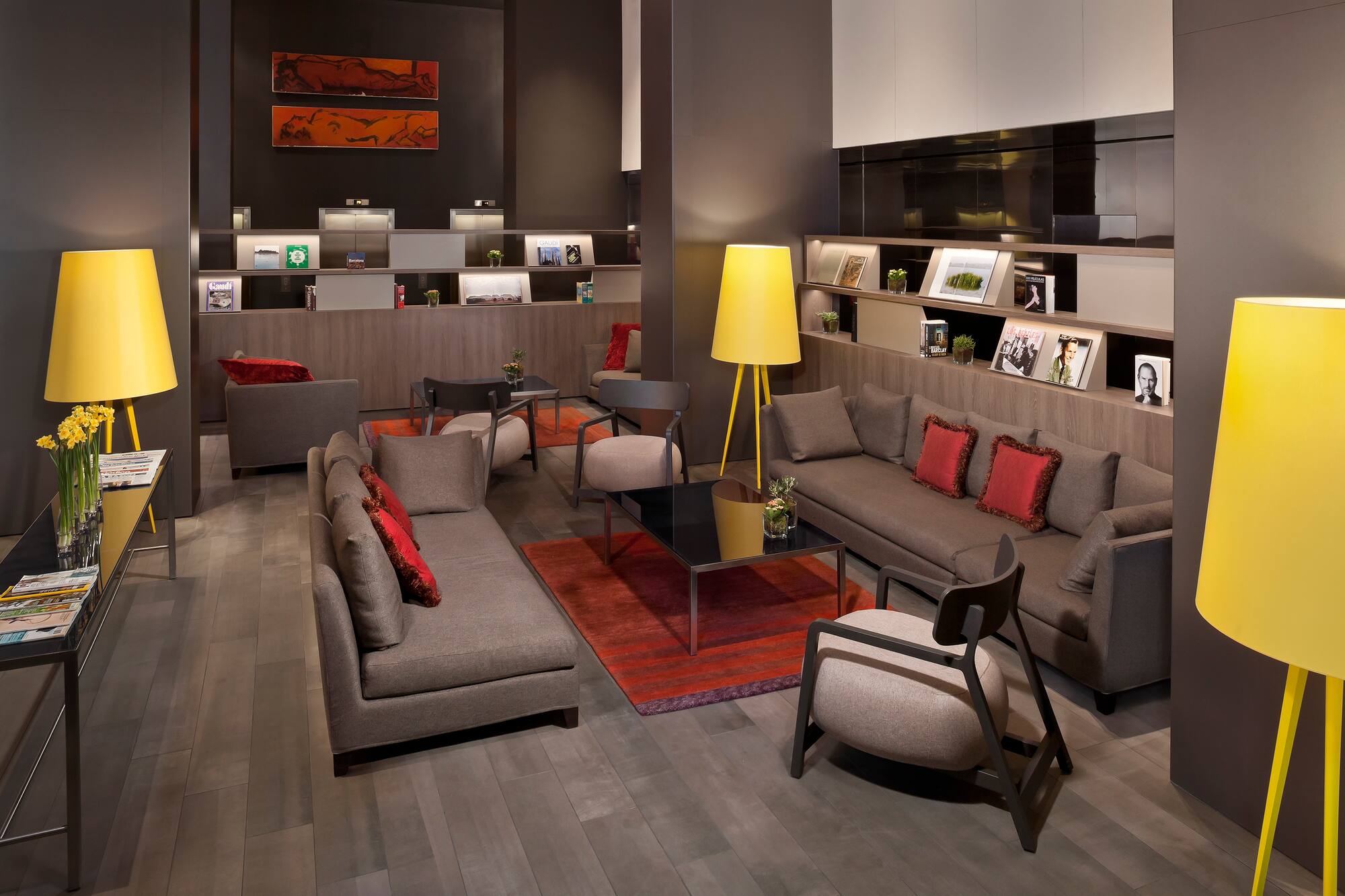 Modern hotel lounge with grey seating, red accents, yellow lamps, and shelving.