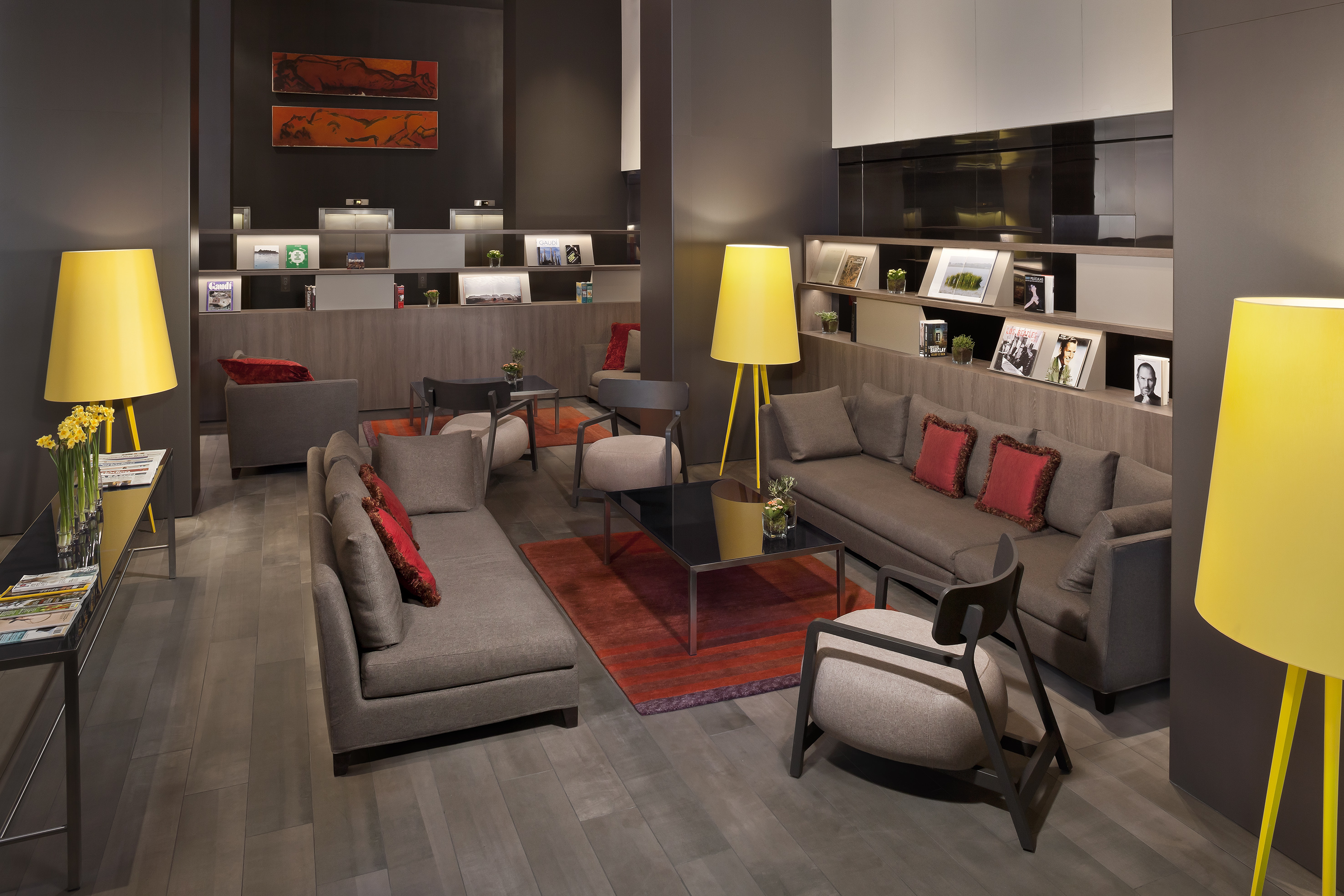 Modern hotel lounge with grey seating, red accents, yellow lamps, and shelving.
