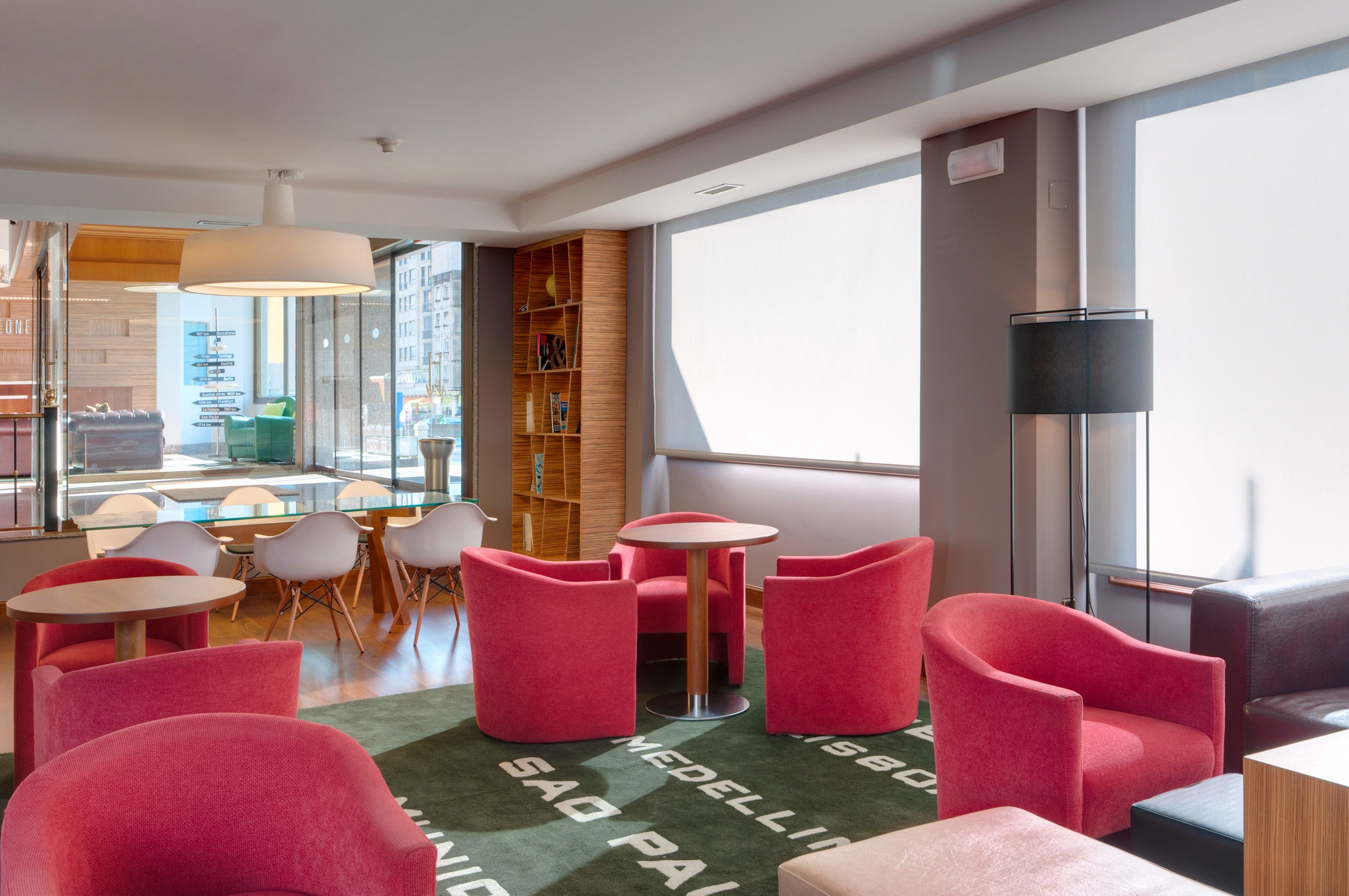 Modern hotel lounge with pink seating, wooden tables, and a green city-name rug.