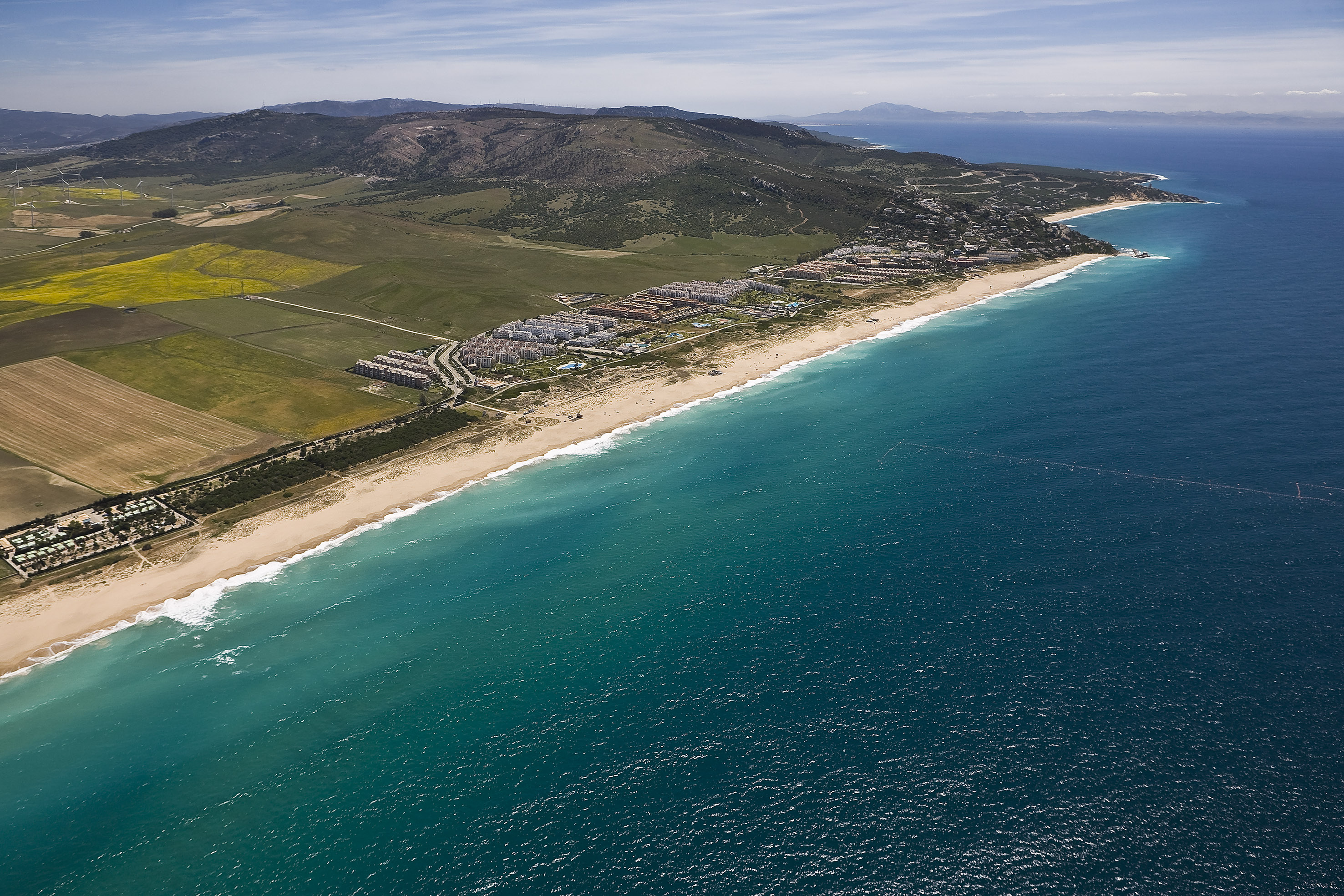 Aerial view: Turquoise beach, coastal resort, green fields, distant mountains.