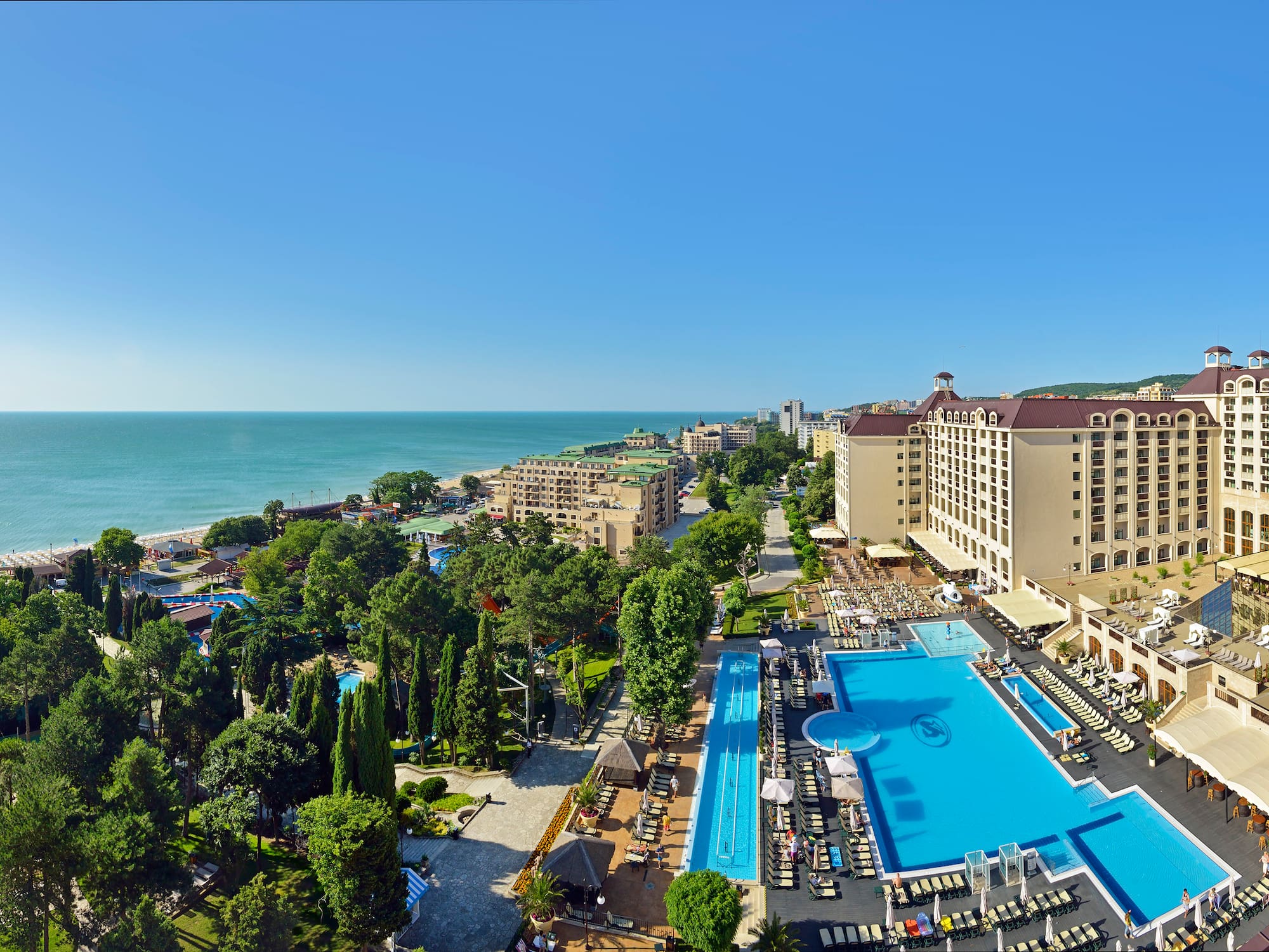 Beachfront resort with large hotel buildings, blue pools, lush gardens, and sea.