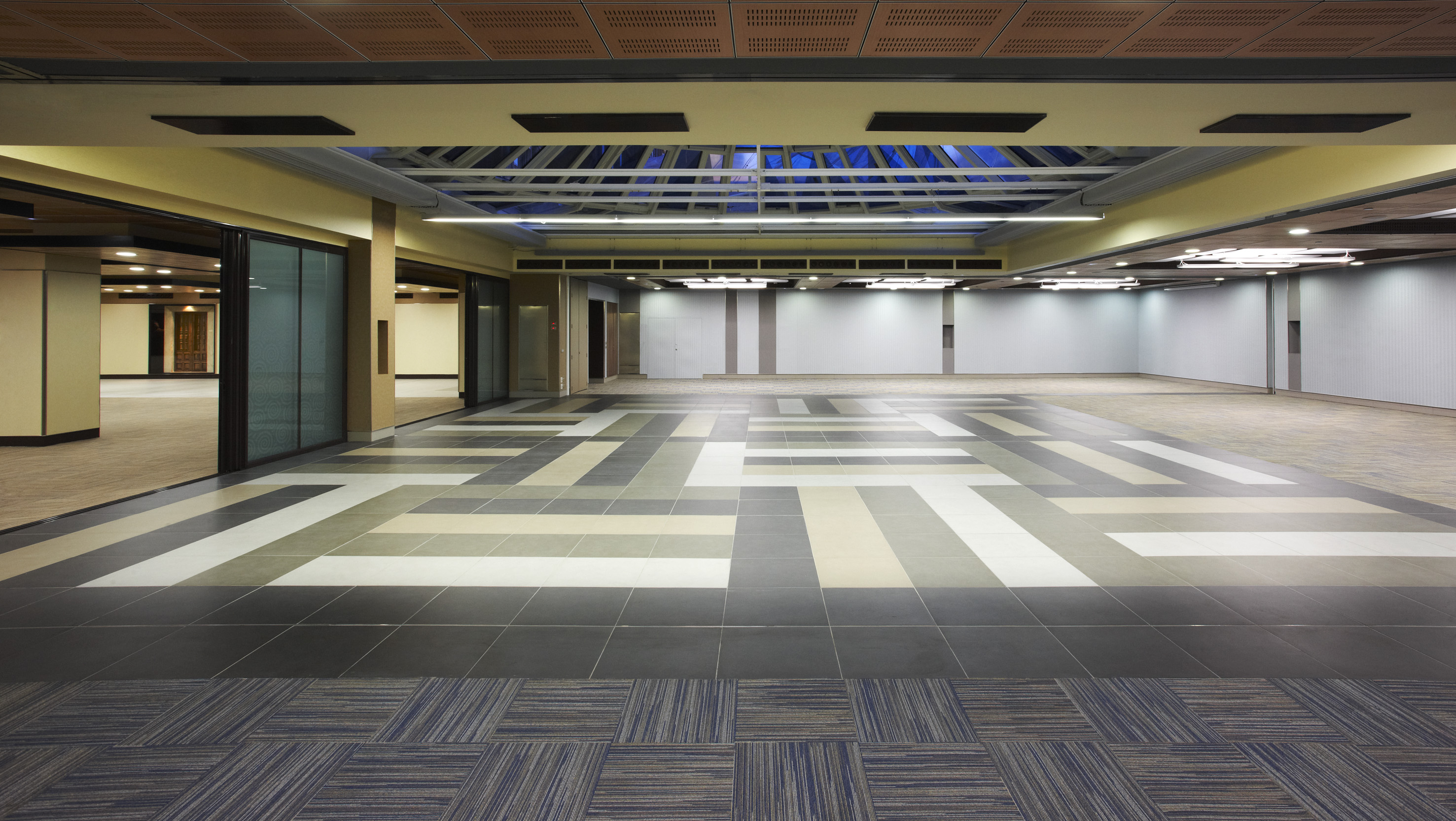 Empty modern hall with geometric floor tiles, ceiling skylight, and glass doors.