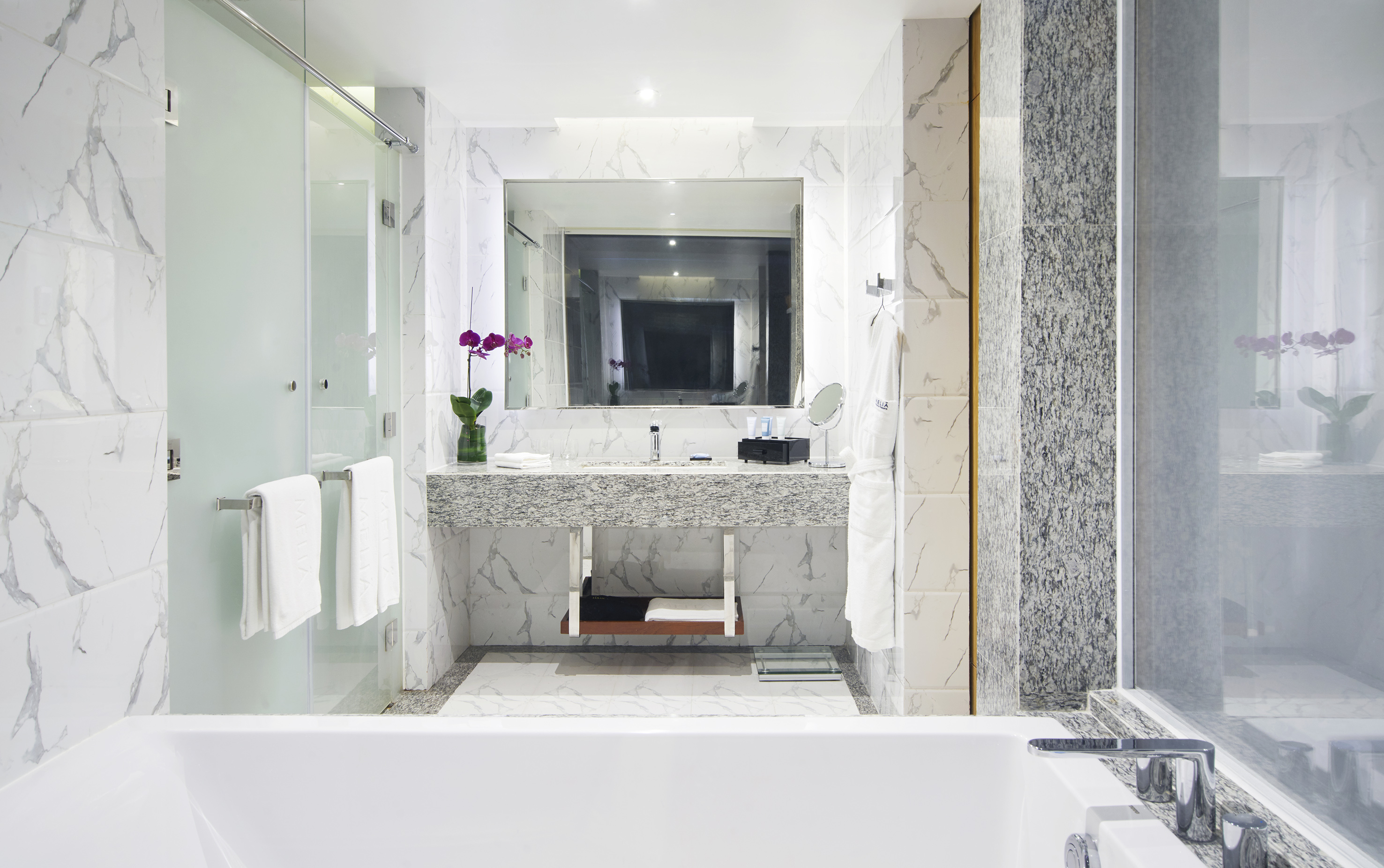 Luxury hotel bathroom with marble walls, bathtub, and glass shower.