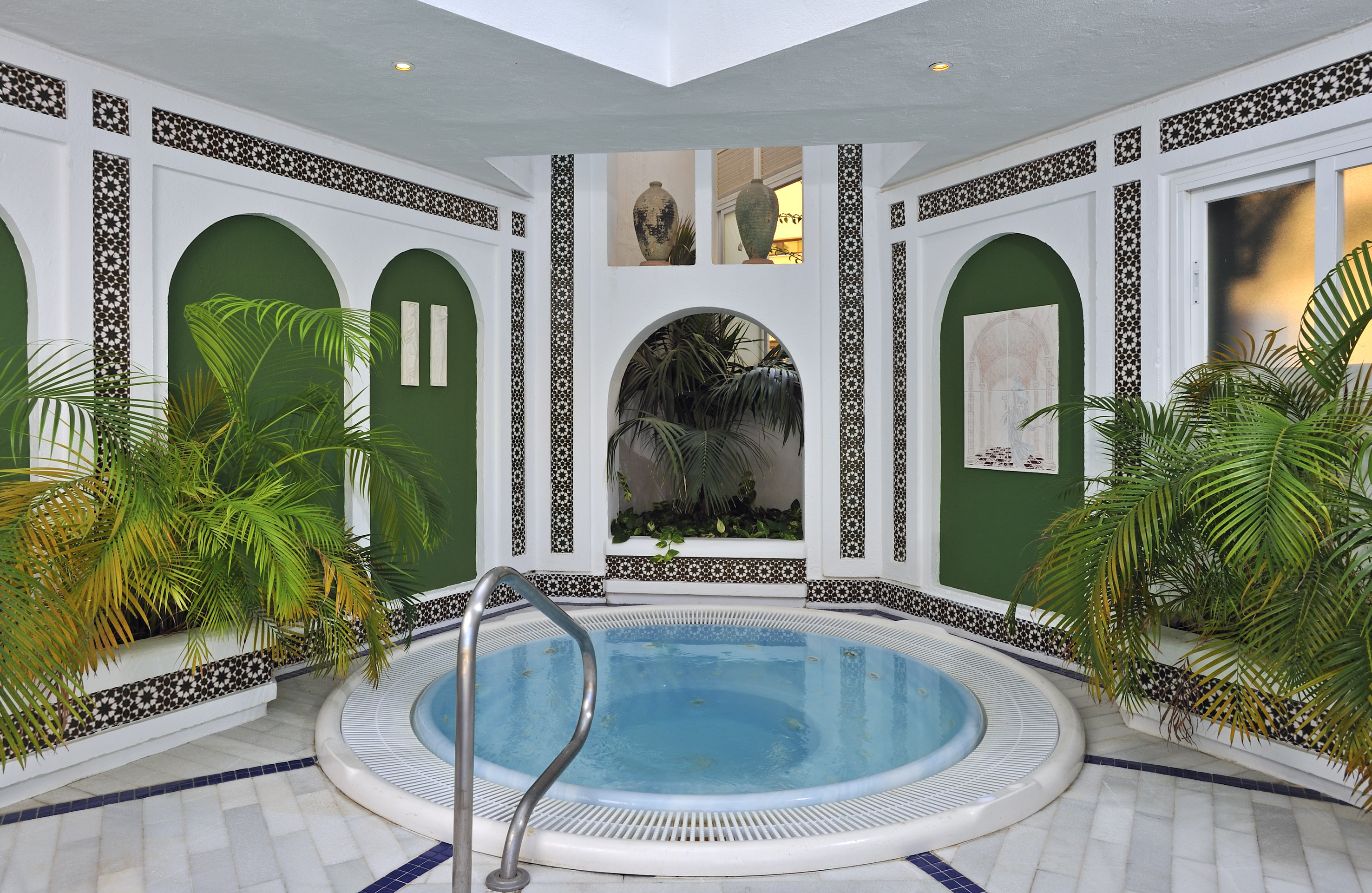 Indoor spa hot tub with Moroccan tile, green arched walls, and tropical plants.