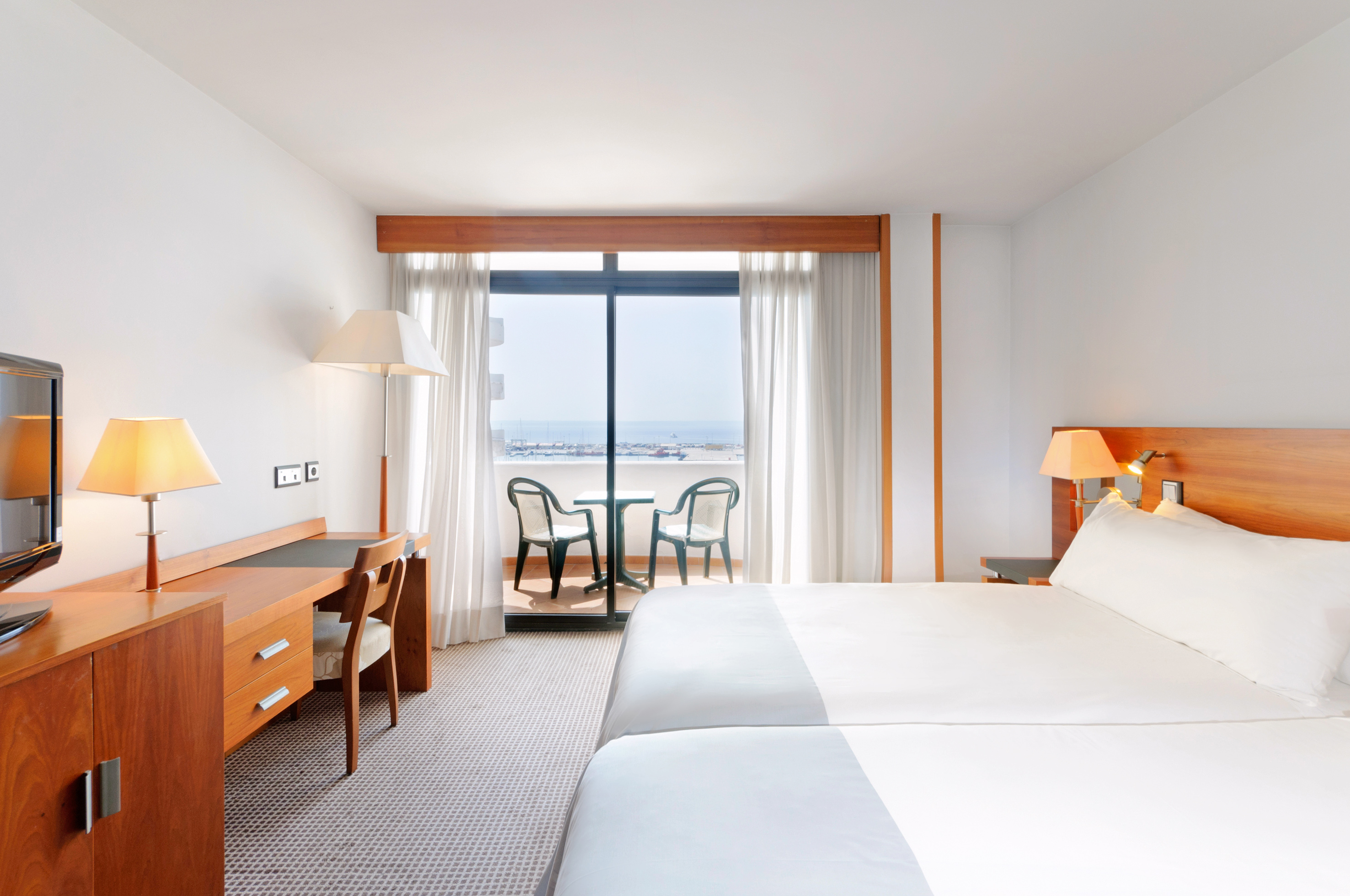 Bright hotel room with twin beds, workspace, and balcony overlooking the sea.