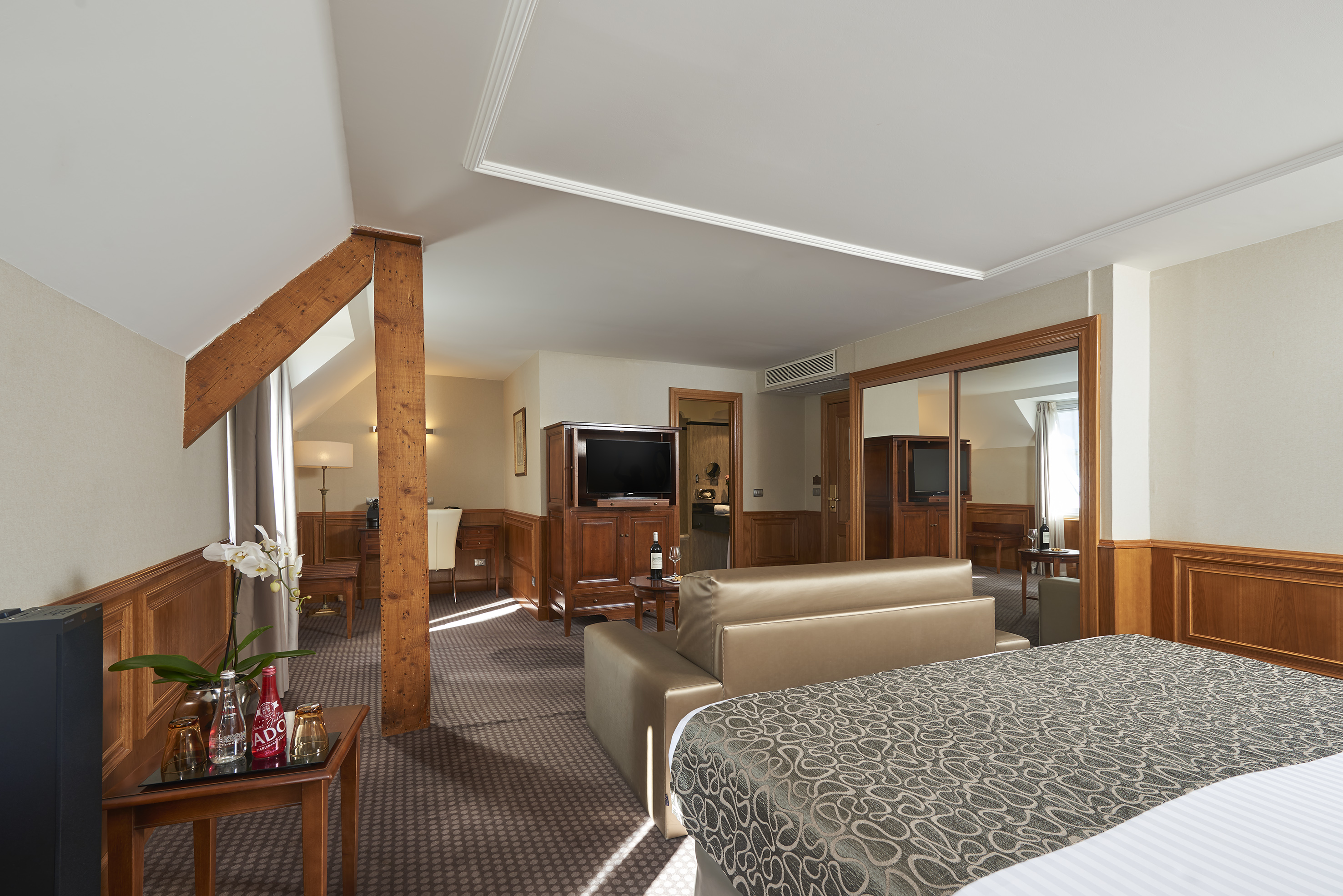 Elegant hotel suite with large bed, sofa, TV, and exposed wooden beams.