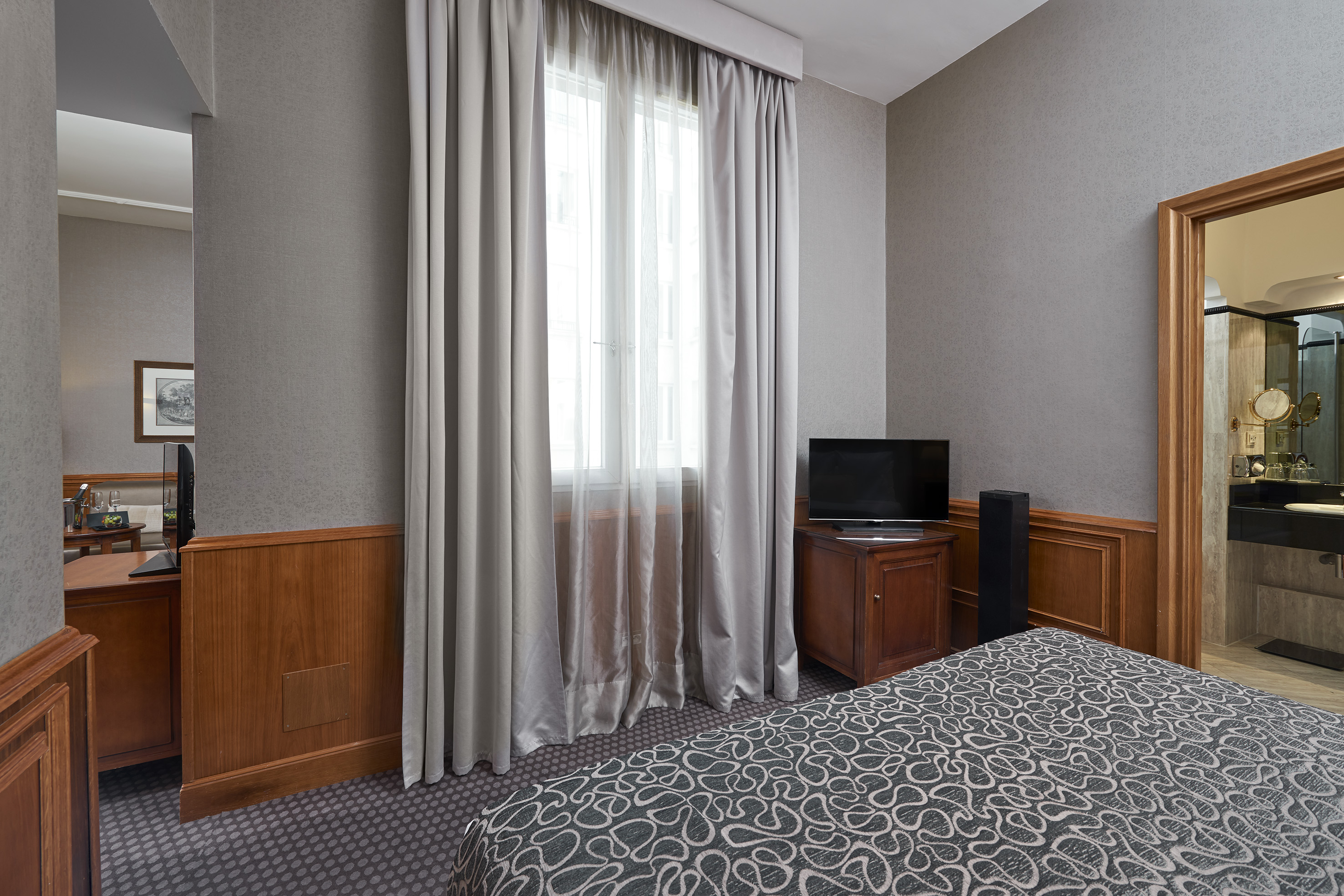 Hotel room with gray walls, patterned bed, window, TV, and wooden accents.