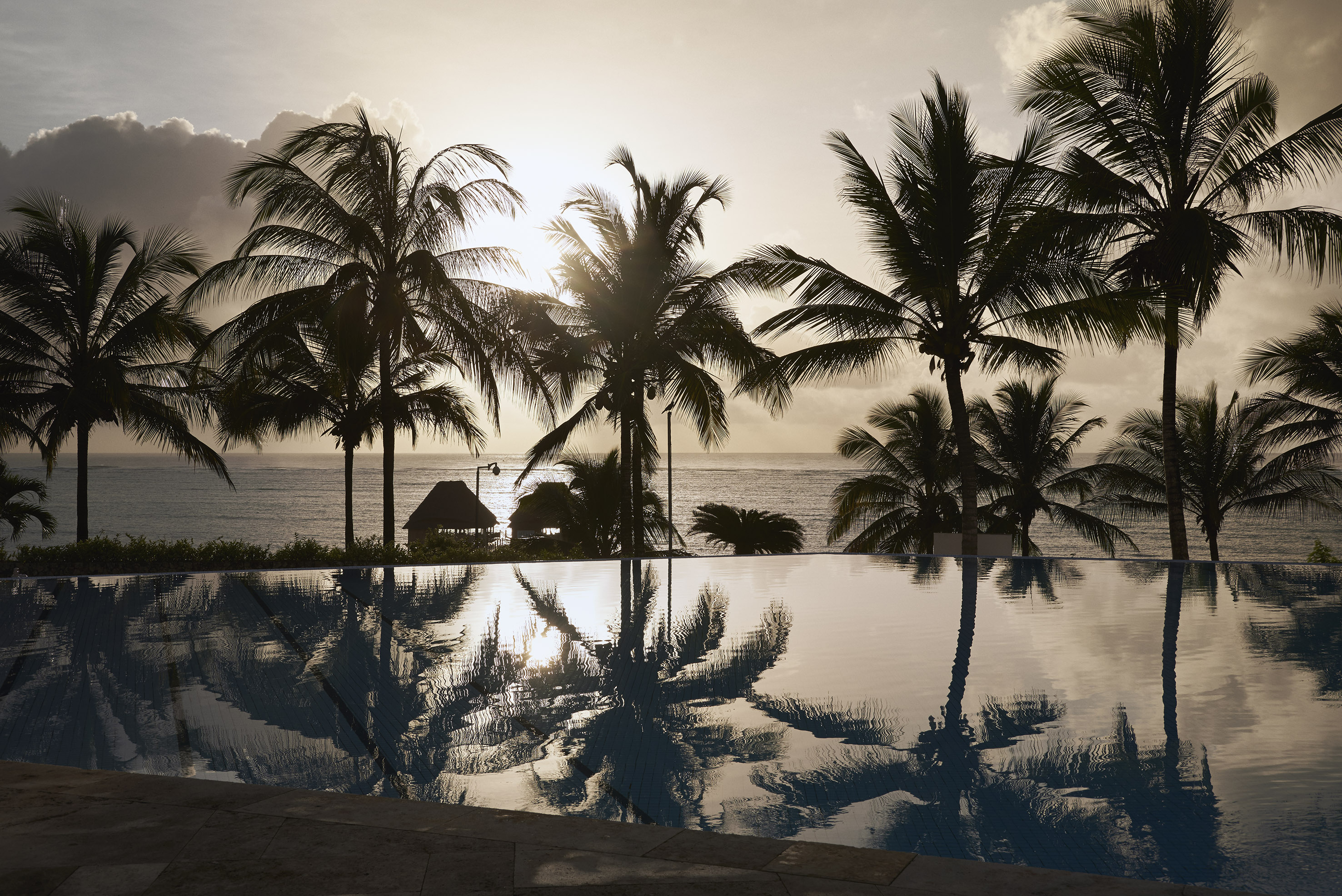 Sunset over tropical ocean, silhouetted palm trees reflect in infinity pool.