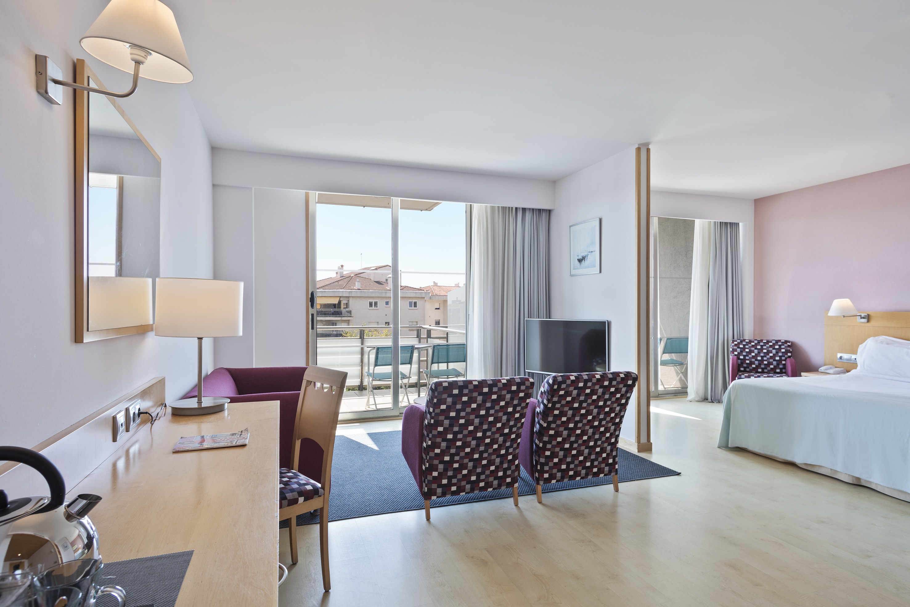 Bright modern hotel suite with living area, bedroom, desk, and balcony.