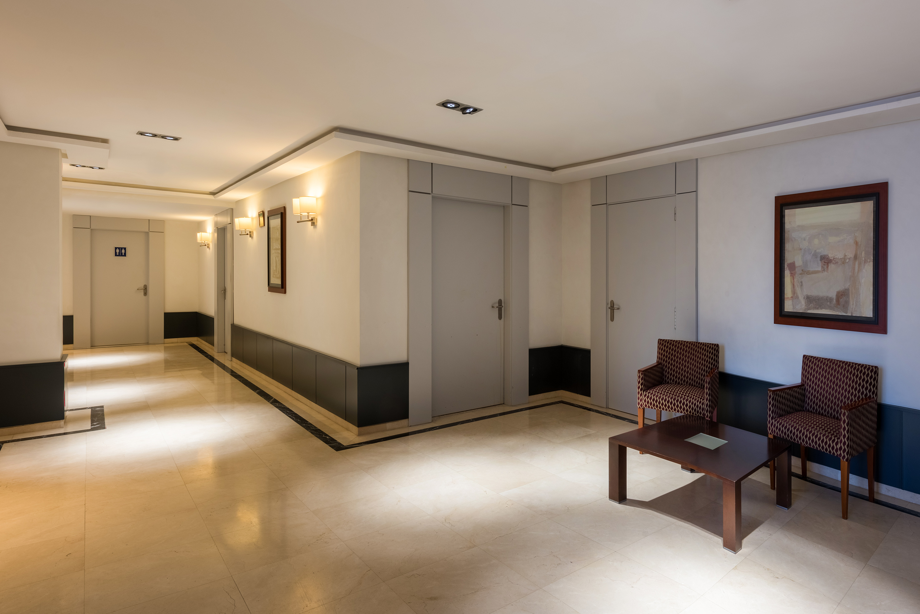 Well-lit building corridor with seating area, tiled floor, doors, and wall art.