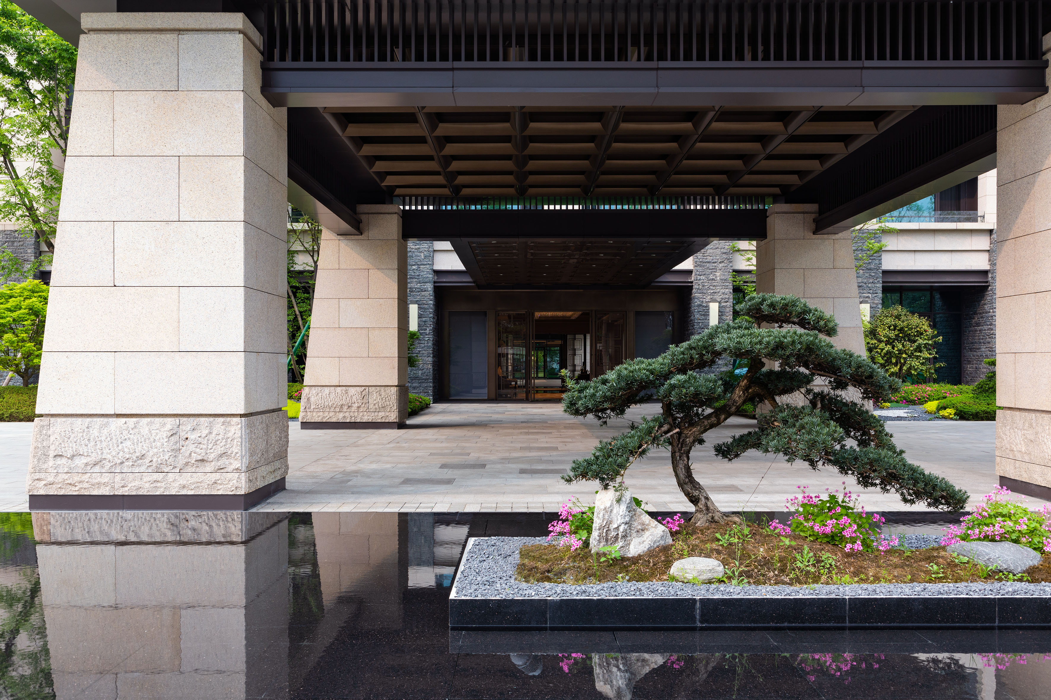 Modern luxury building entrance with stone pillars, water feature, and bonsai tree.