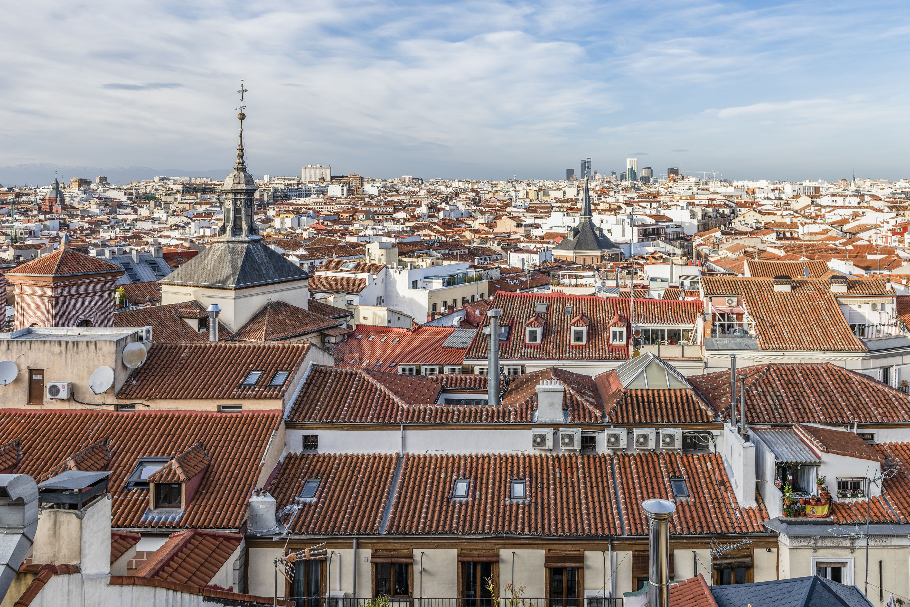 Madrid cityscape with red-tiled rooftops, spires, and distant urban skyline.