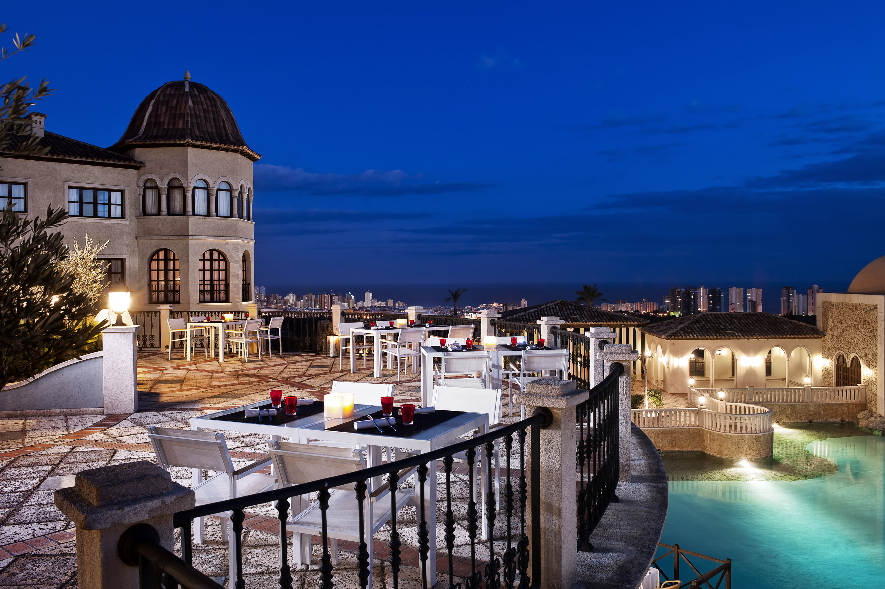 Luxury hotel terrace dining at night with city skyline and illuminated pool.