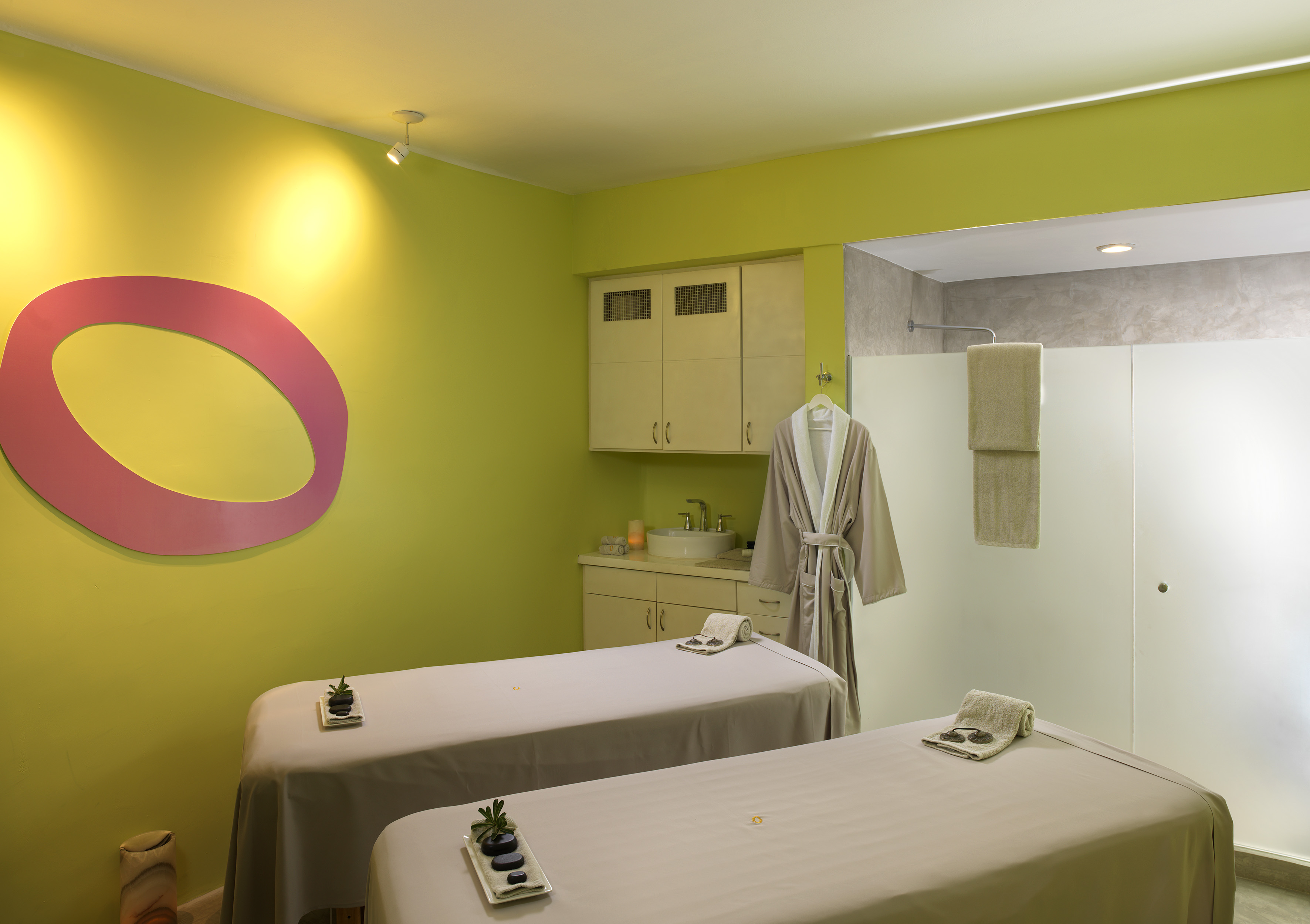 Green spa treatment room with two massage beds, hanging robe, and shower.