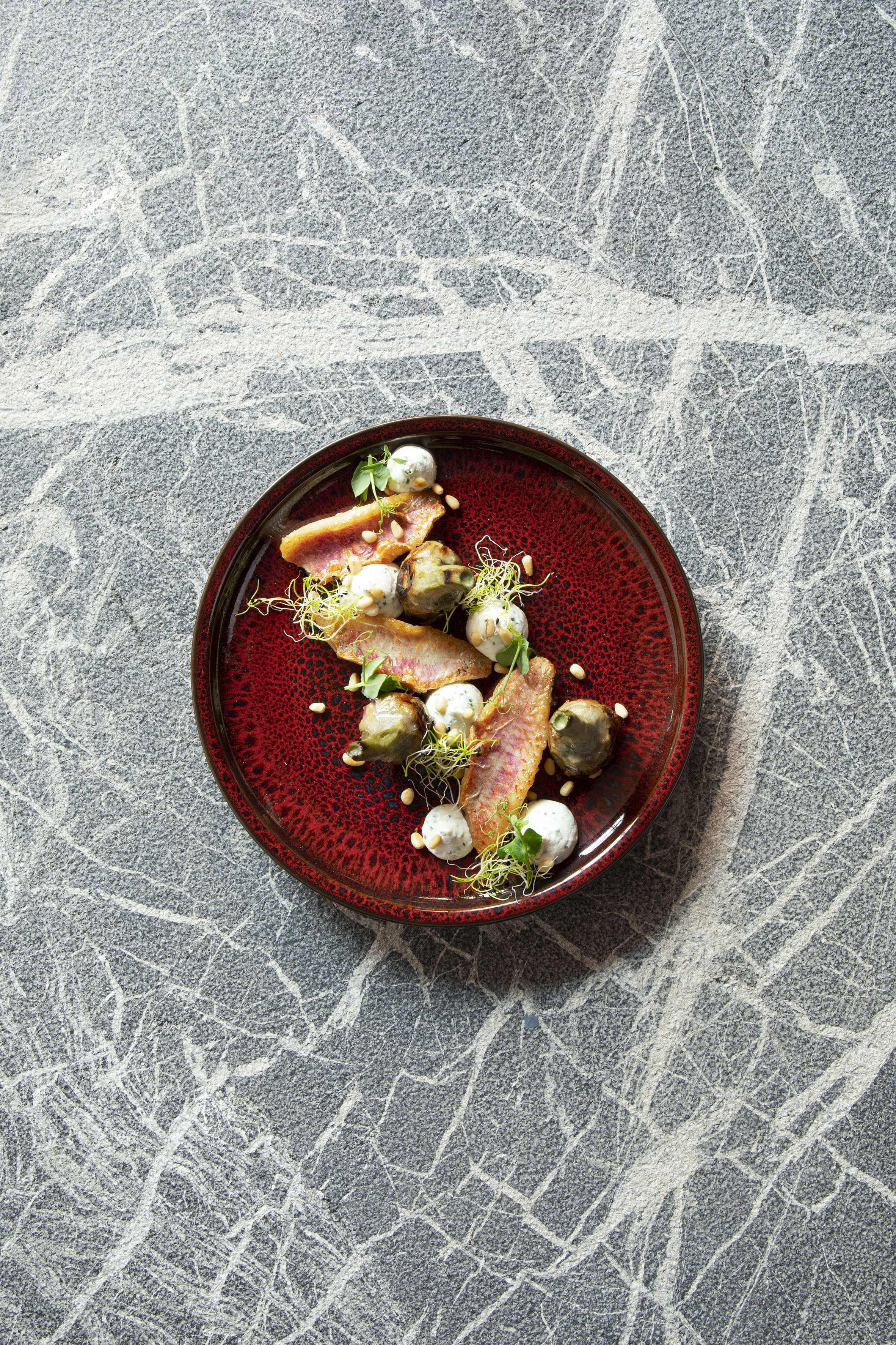 Red mullet fish with artichokes, sprouts, and pine nuts on a red plate.