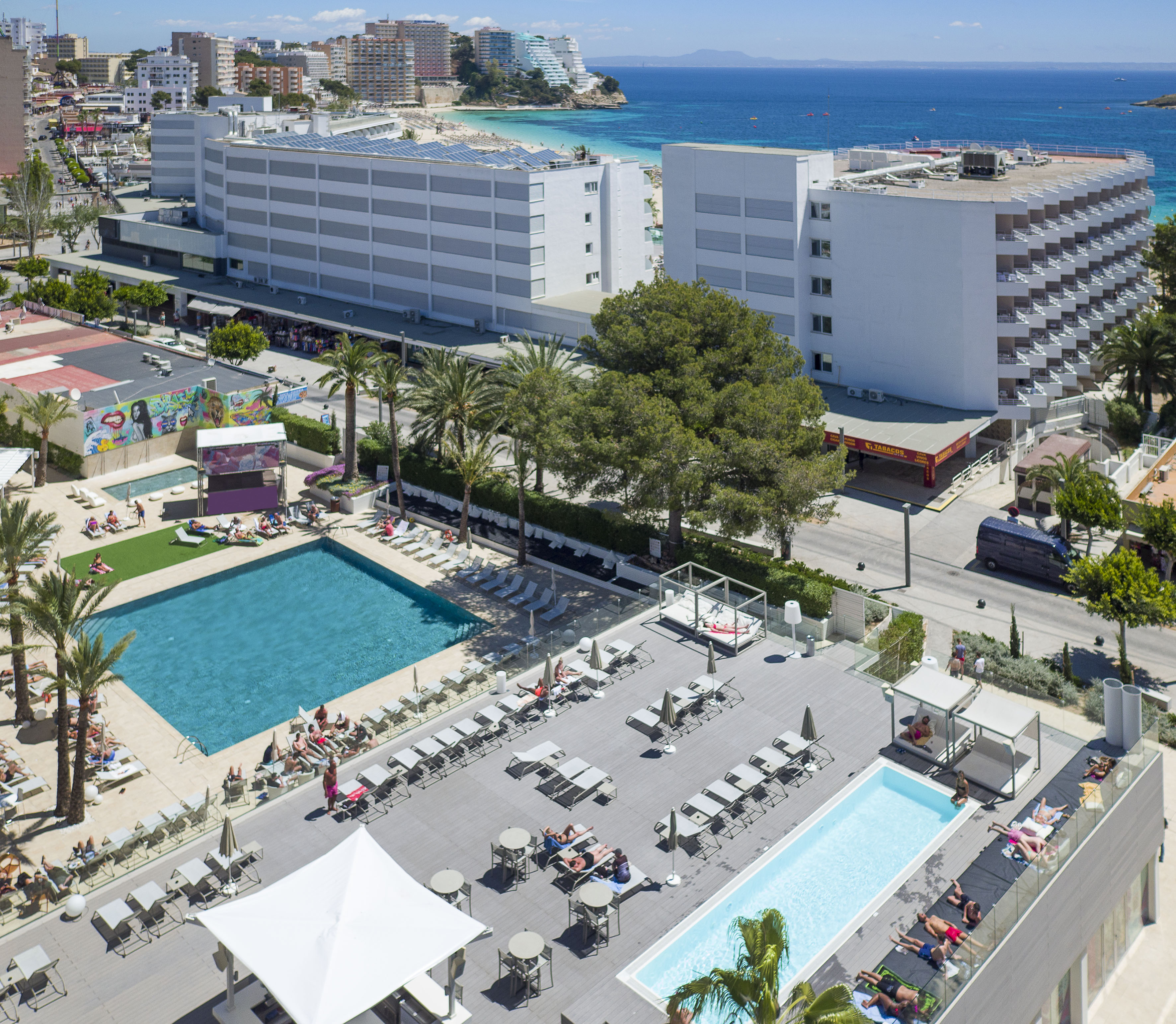 Coastal resort with pools, sunbathers, palm trees, hotels, and blue sea.