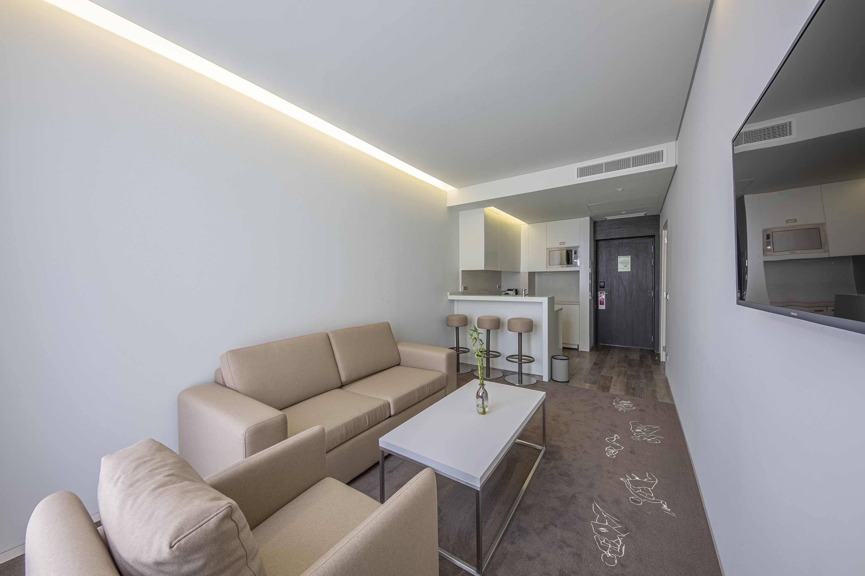 Modern hotel living room with beige sofa, white coffee table, and kitchenette.