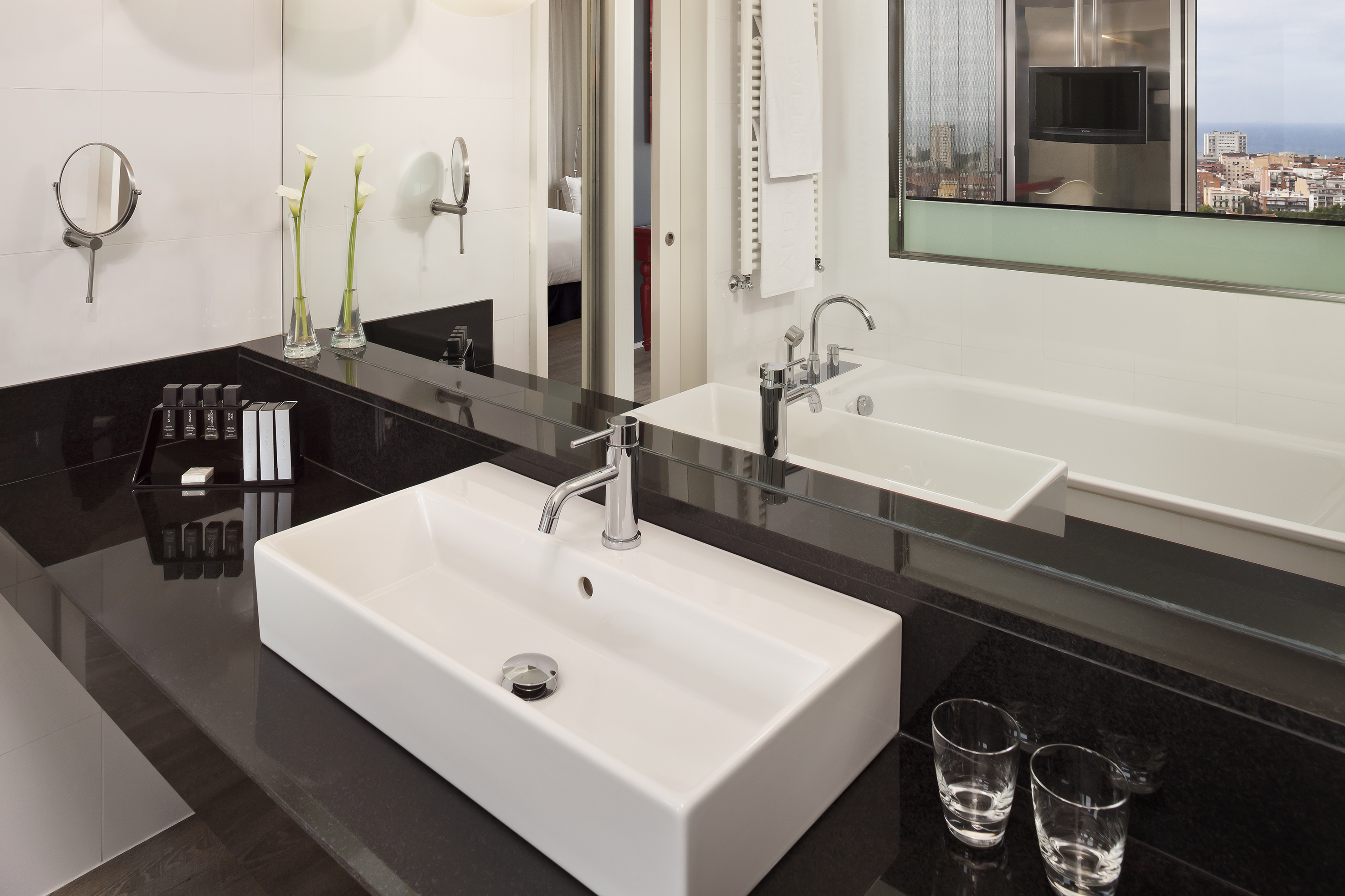Modern hotel bathroom with white sink, black counter, bathtub, and city view.