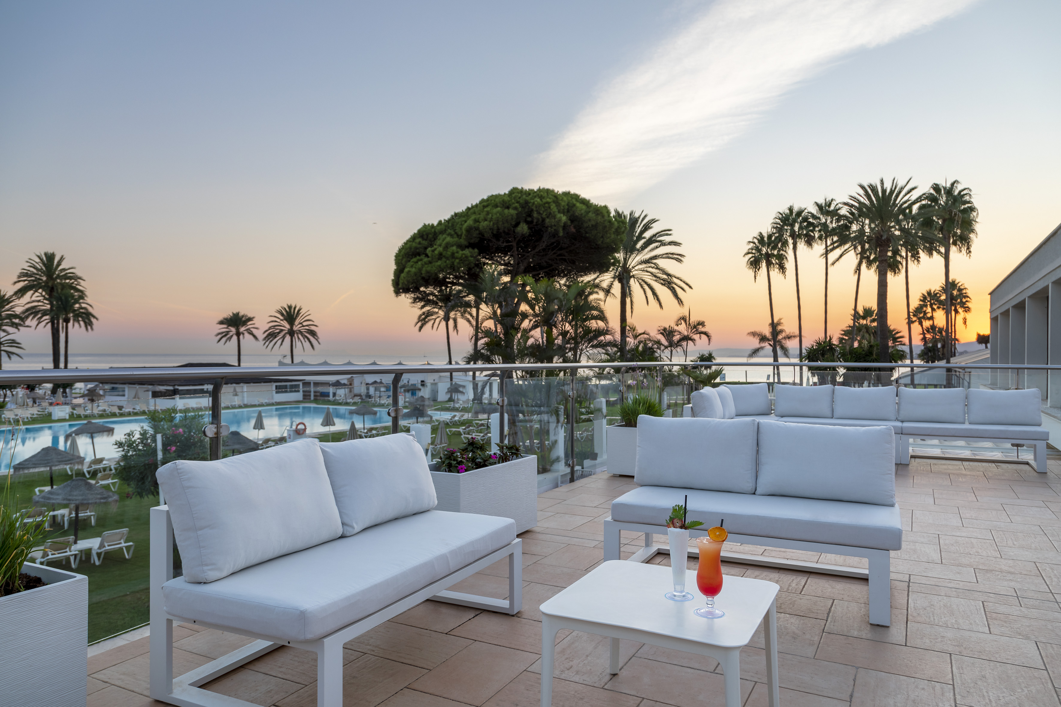 Hotel terrace with white sofas, cocktails, palm trees, ocean view at sunset.