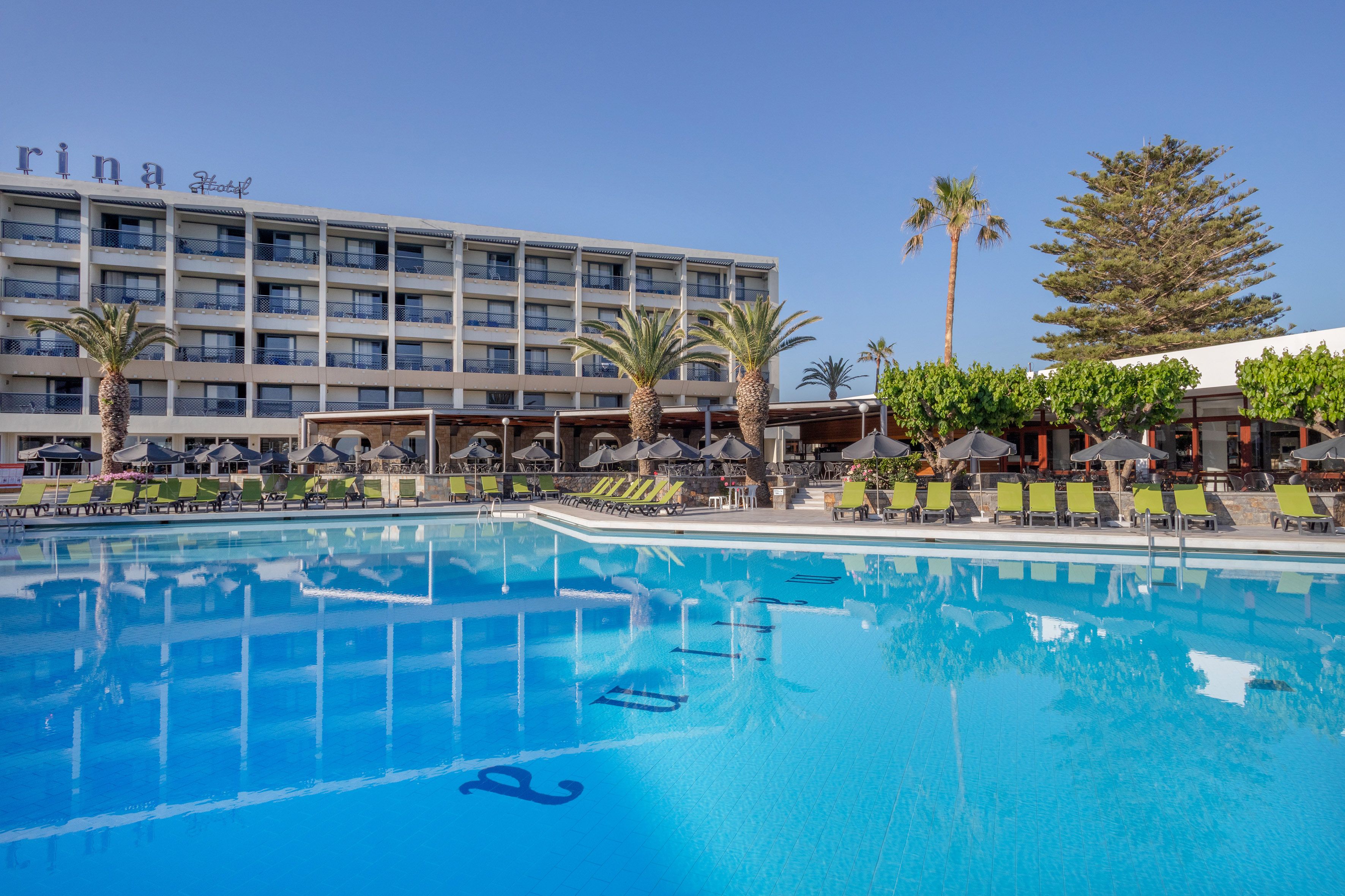 Hotel swimming pool with green sun loungers, palm trees, and resort building.