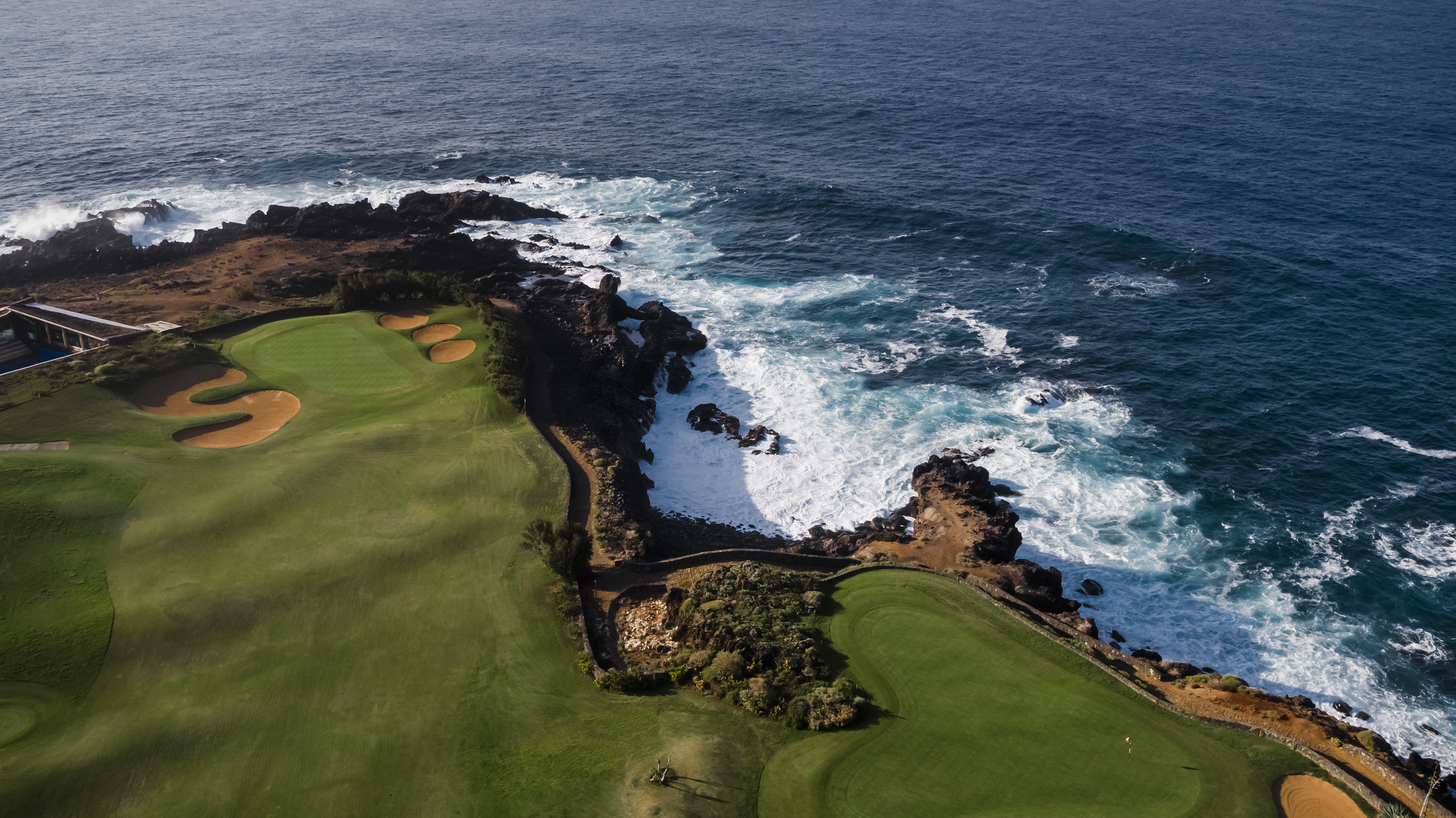 Aerial oceanfront golf course, green fairways, sand bunkers, crashing waves.