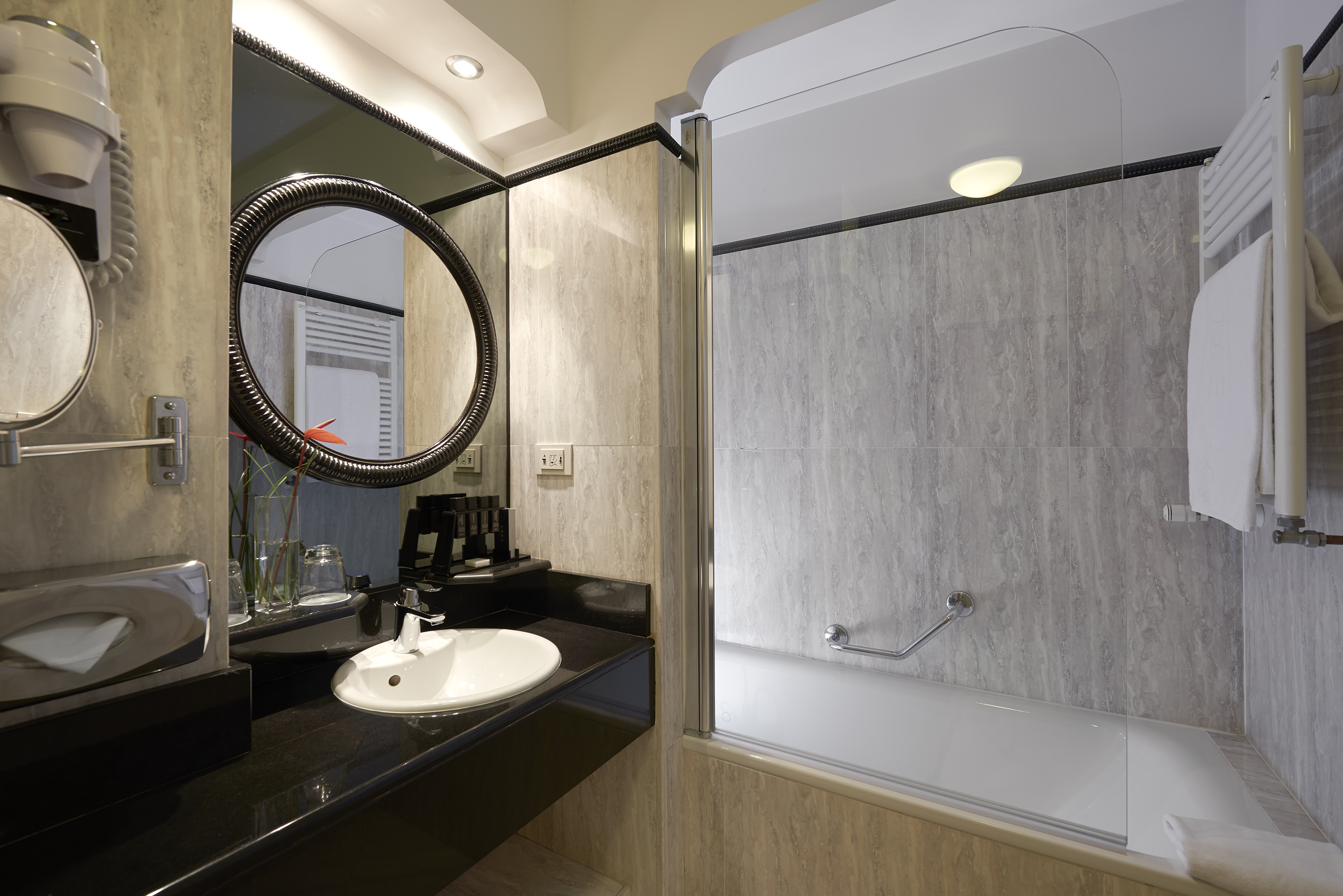 Hotel bathroom vanity, white sink, round mirror, and bathtub with tiled walls.