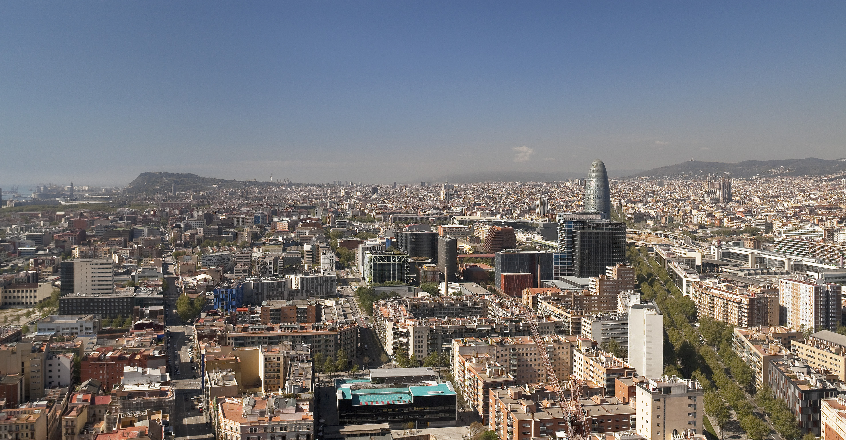Vista aérea de Barcelona con Torre Glòries, Sagrada Familia y Montjuïc bajo cielo azul.