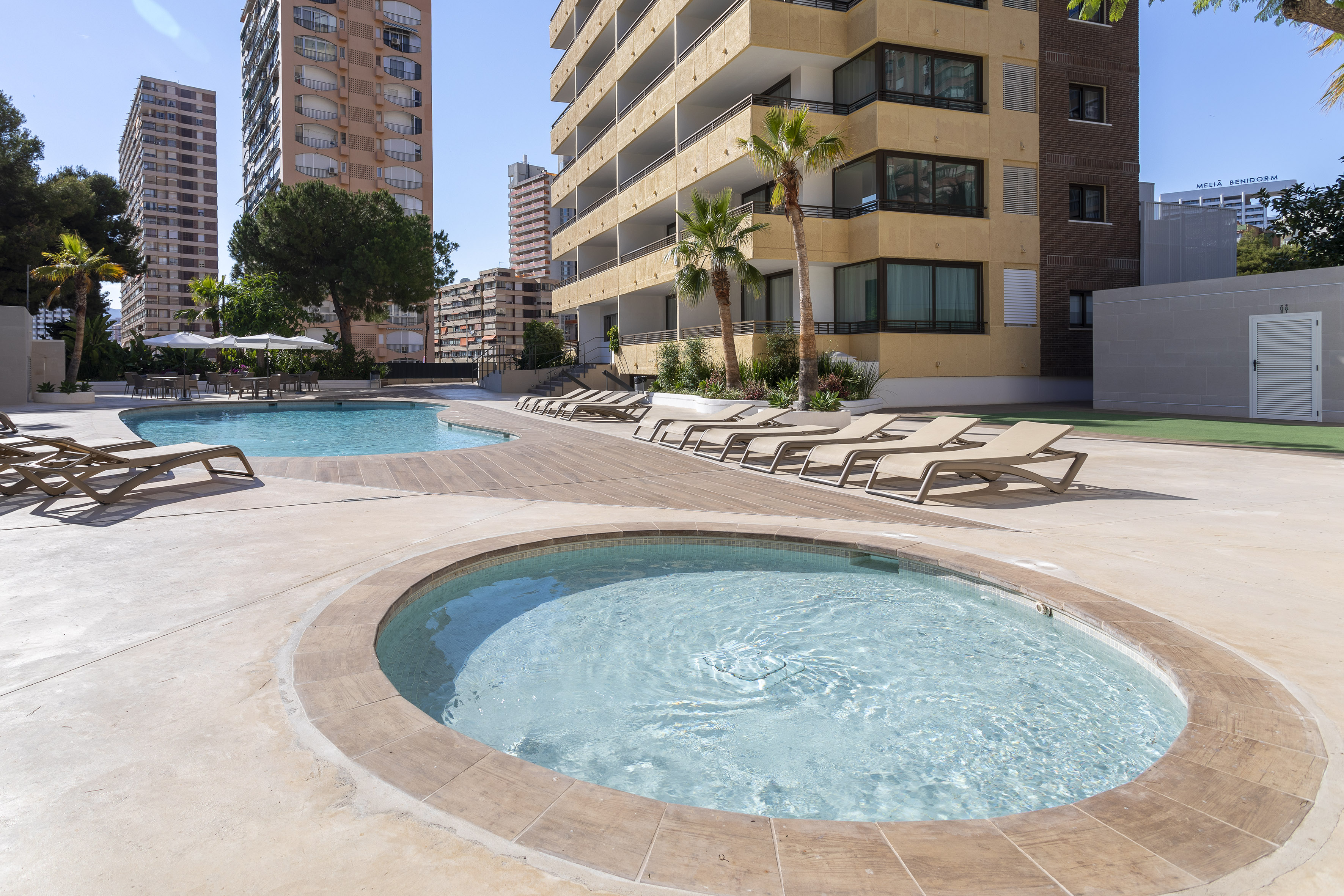 Two swimming pools with sun loungers on a deck, surrounded by tall buildings.