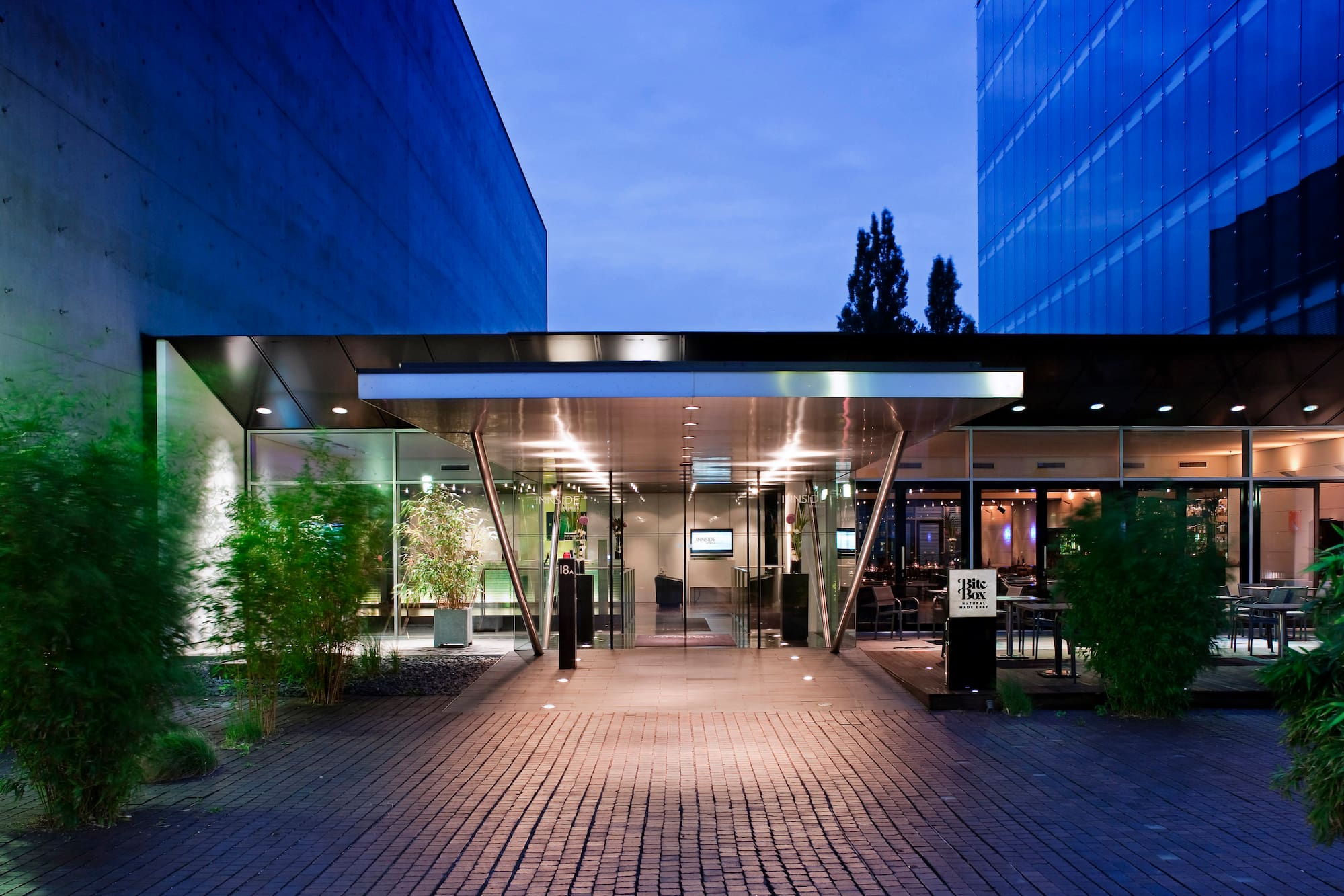 Illuminated modern hotel entrance, glass facade, bamboo plants at dusk.