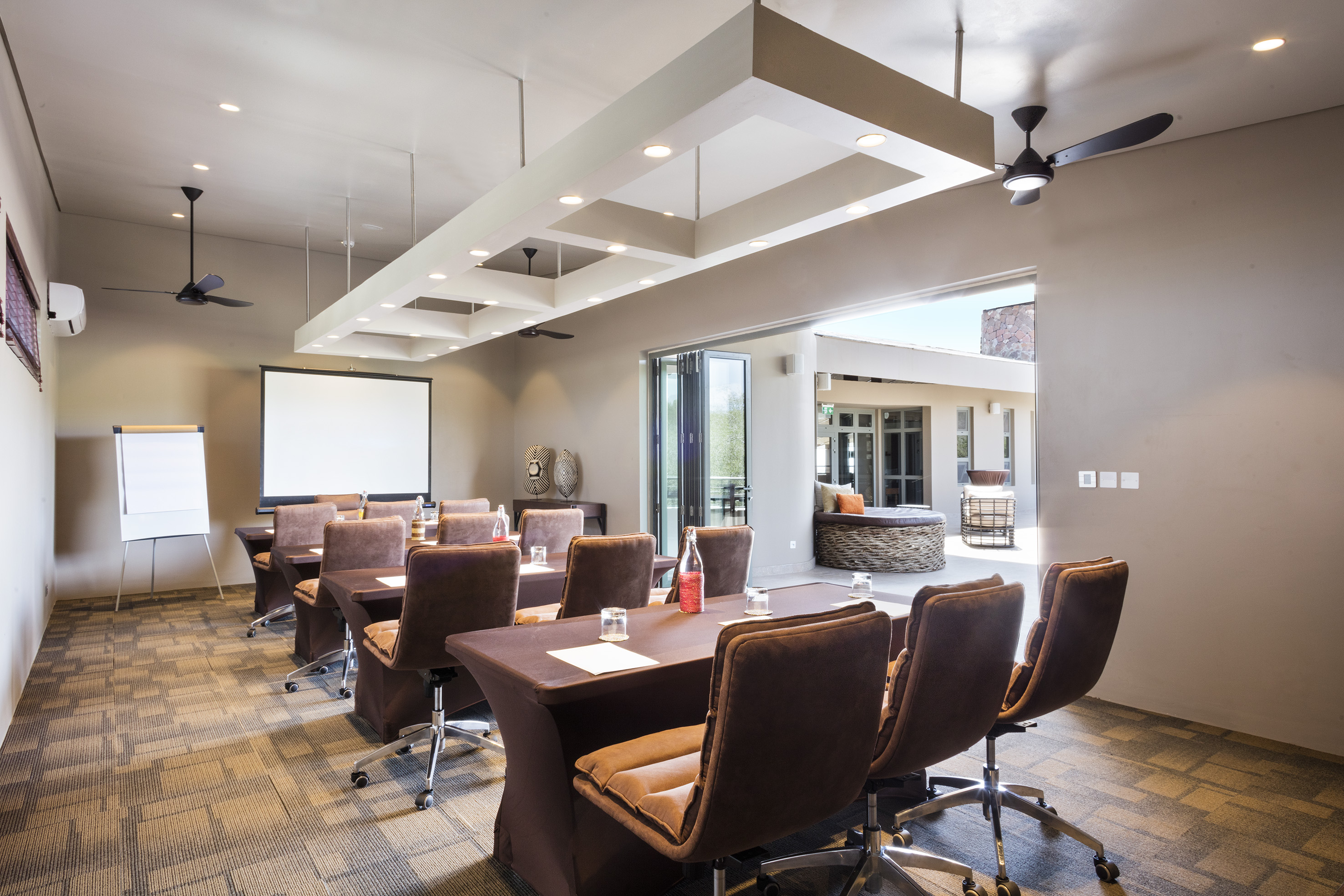 Modern conference room with projector screen, brown tables, and outdoor patio view.