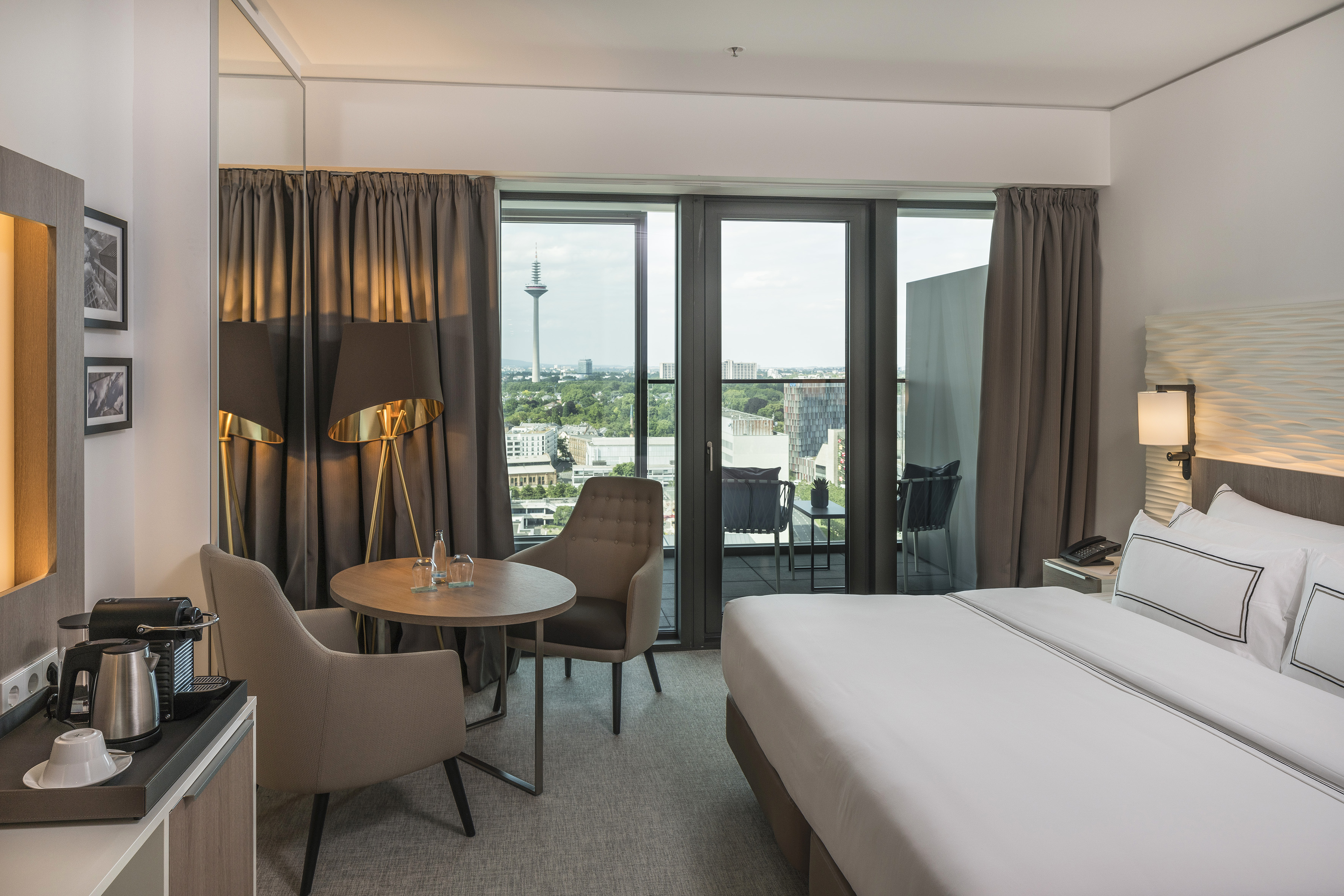 Modern hotel room with Frankfurt city view, balcony, bed, and coffee station.