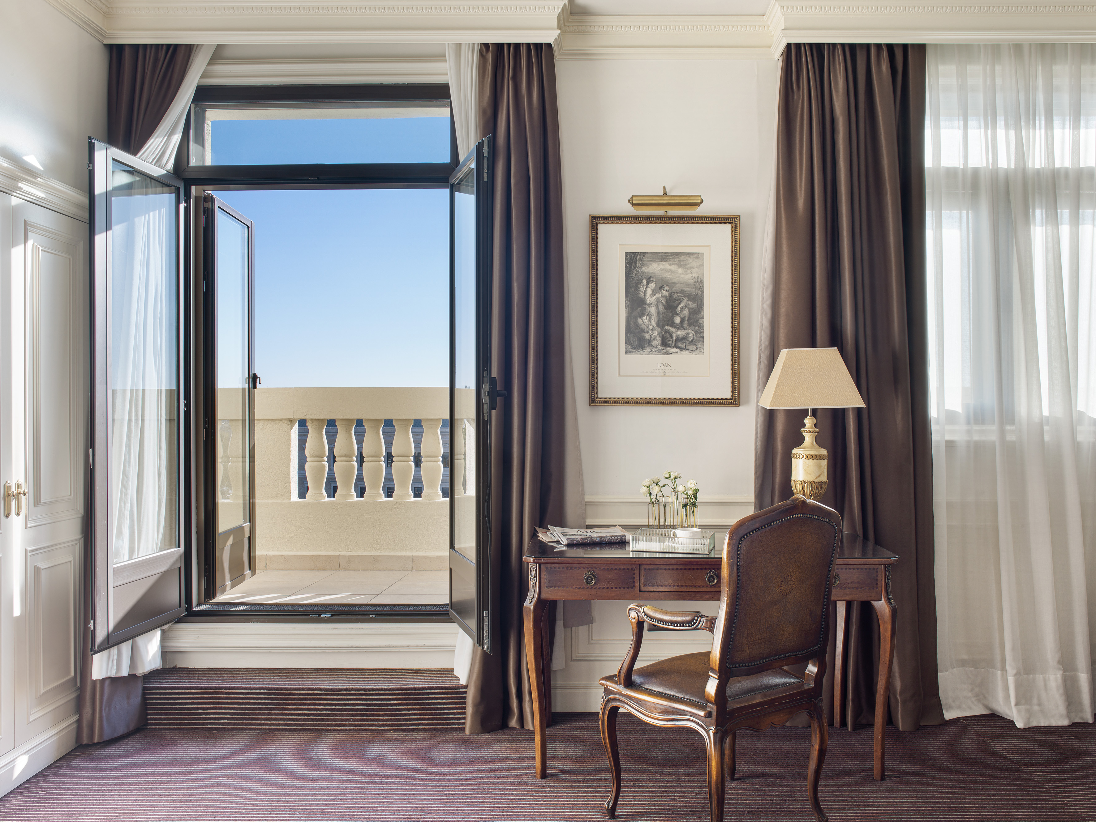 Luxury hotel room with open balcony, desk, leather chair, and classic decor.
