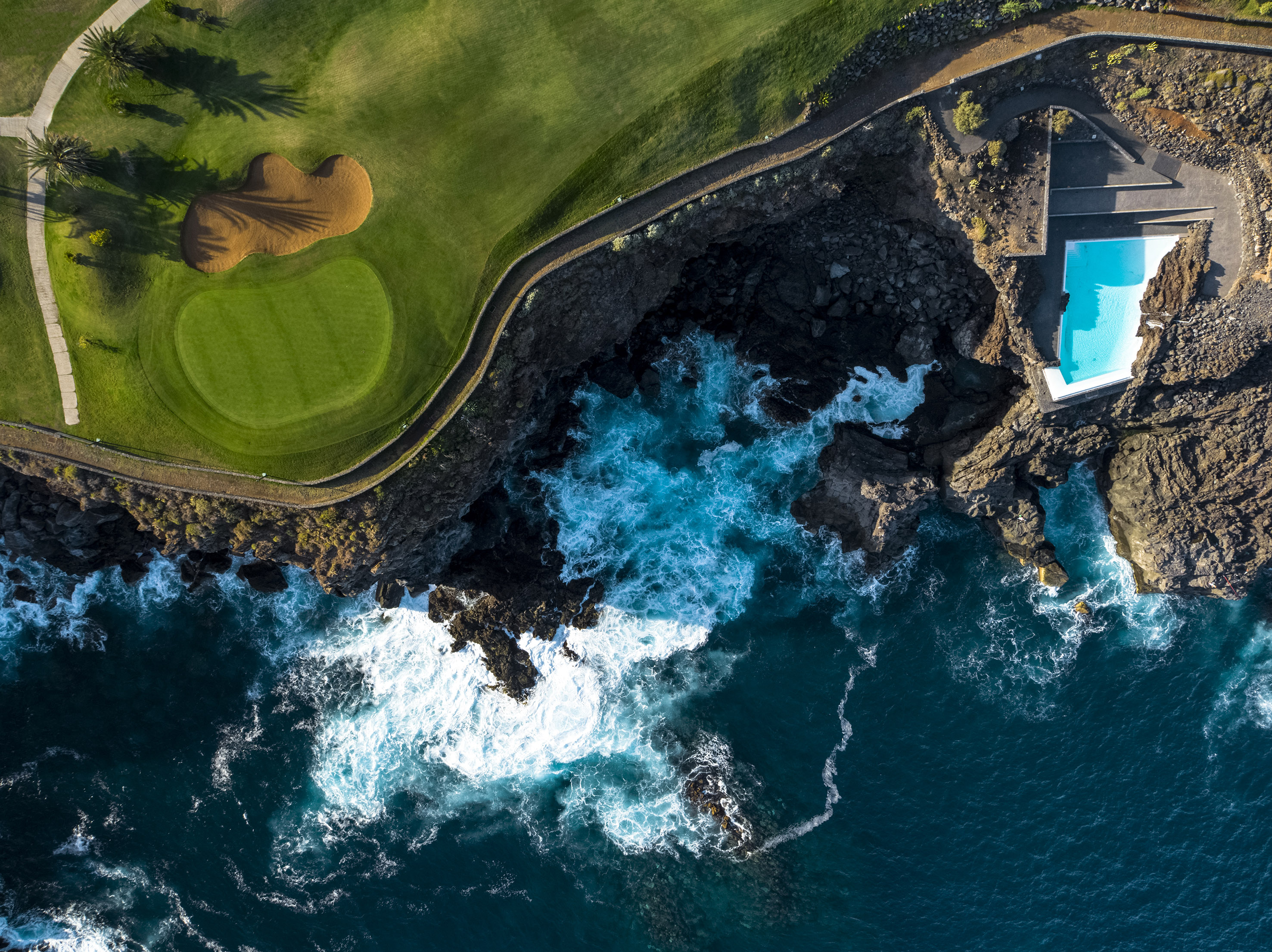 Coastal golf course, rocky cliffs, and cliffside pool by crashing ocean waves.