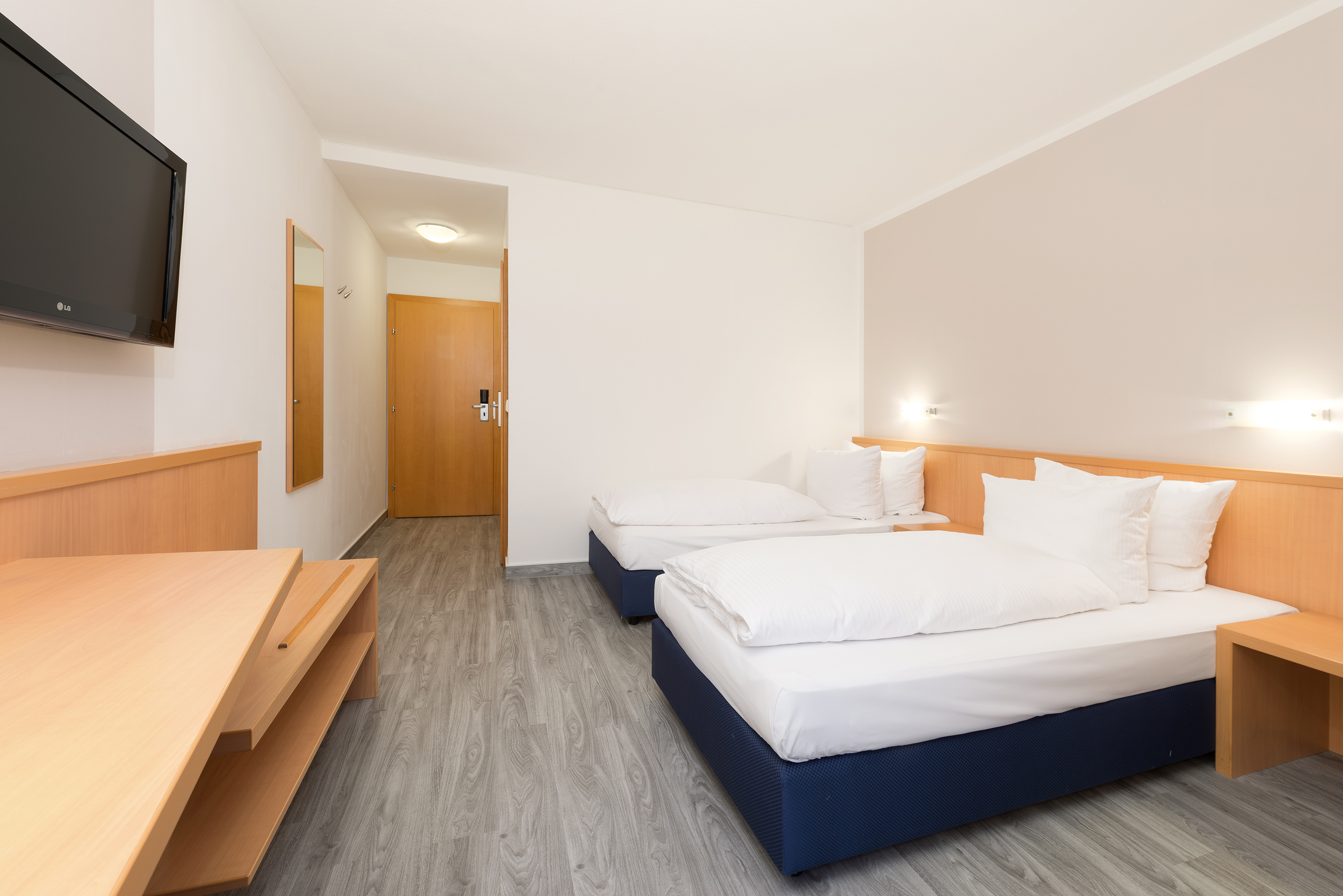 Hotel room with two single beds, wooden desk, wall-mounted TV, and wood floor.