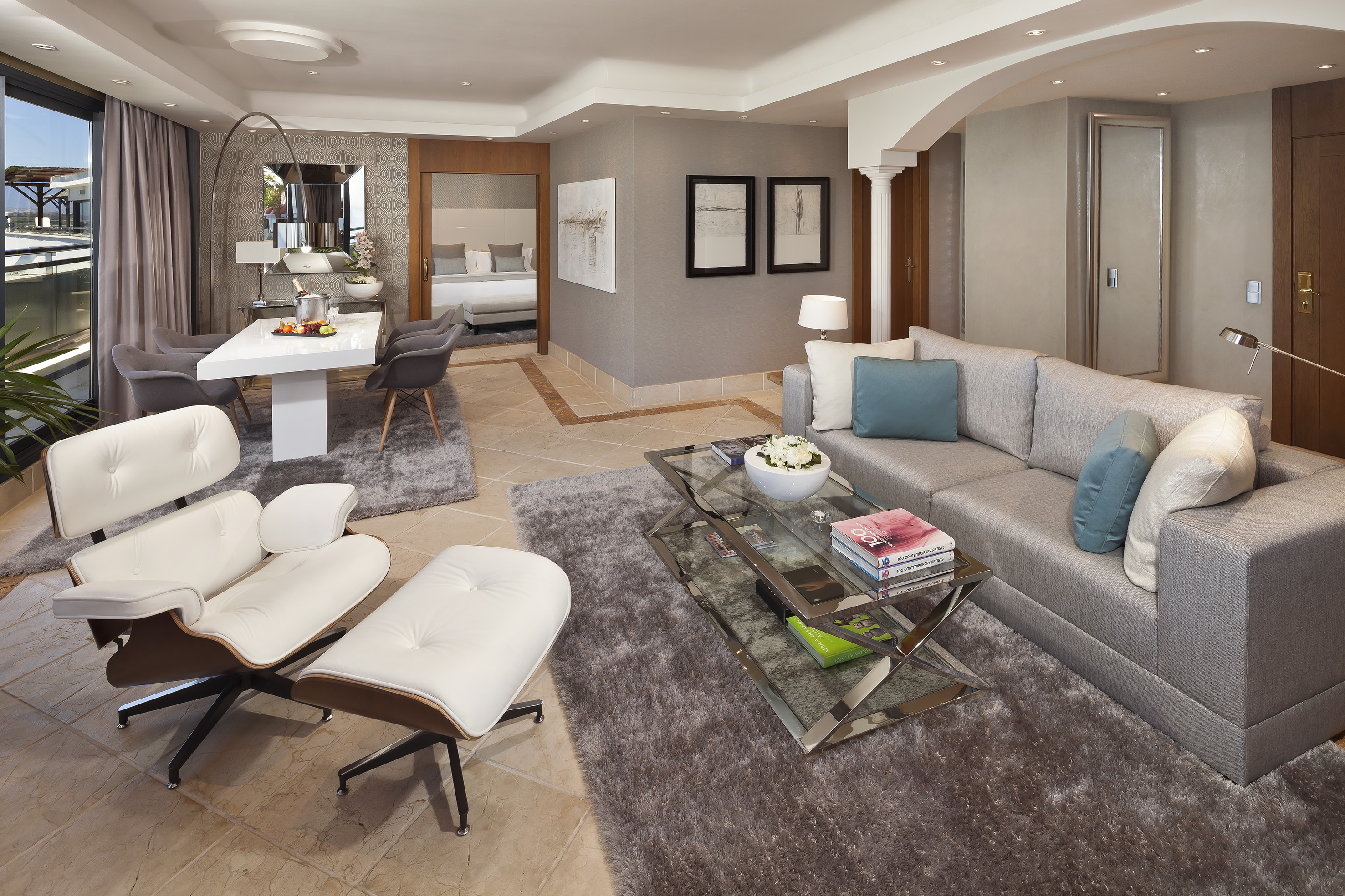 Modern luxury hotel suite living room with grey sofa, Eames chair, and dining area.