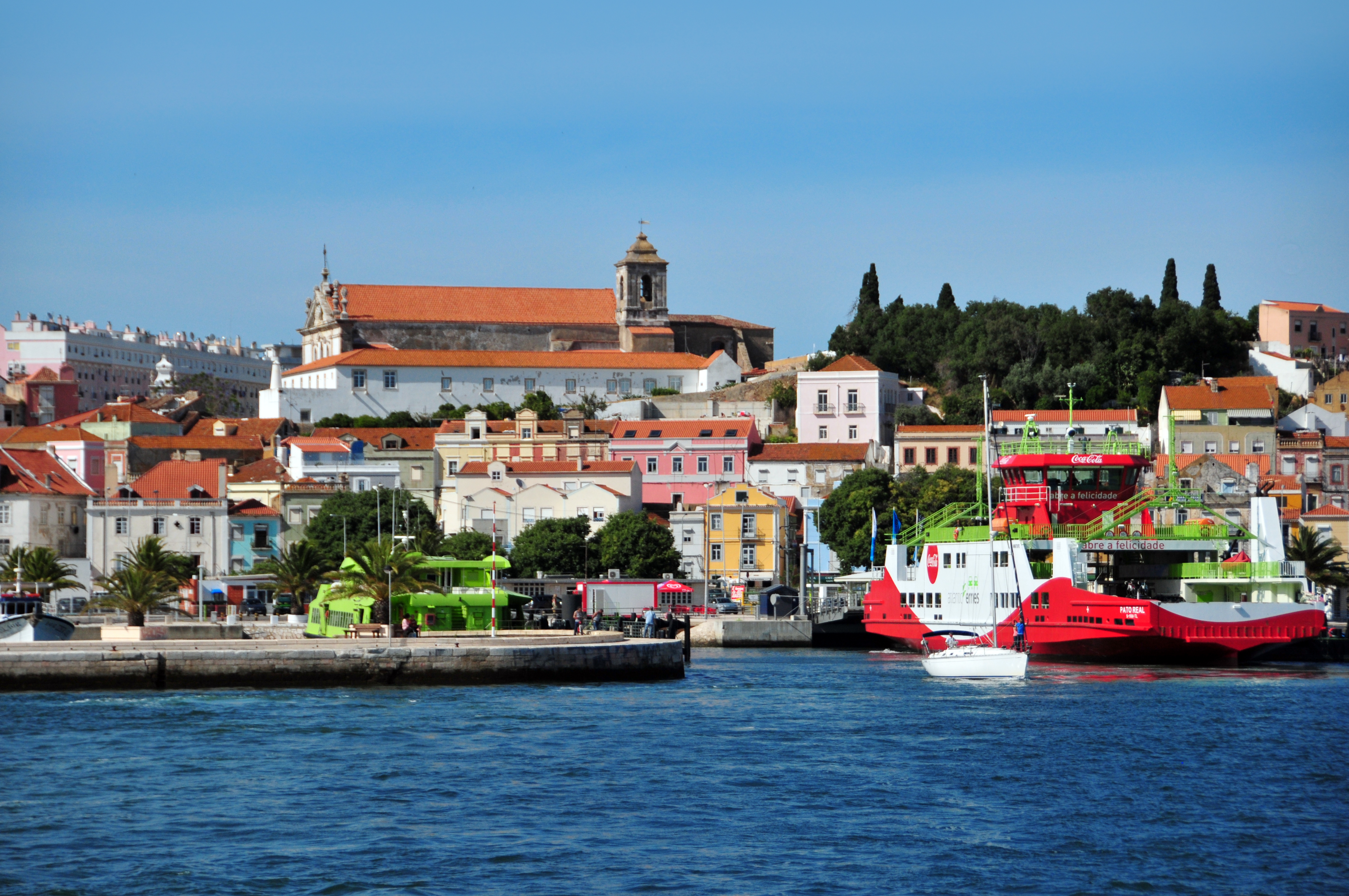 Lisbon waterfront with colorful buildings, church, and a red ferry on blue water.
