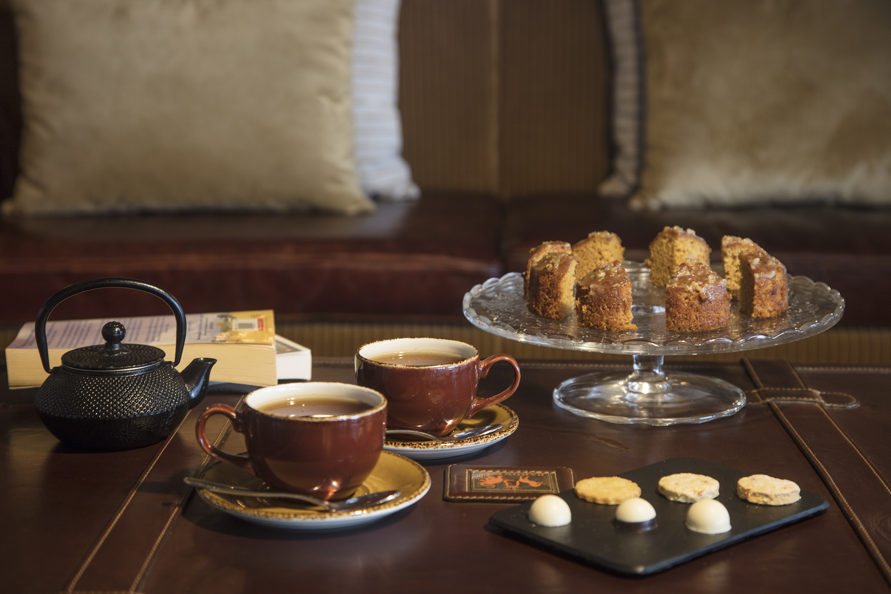 Tea cups, cast iron teapot, cake slices, cookies & chocolates on table.