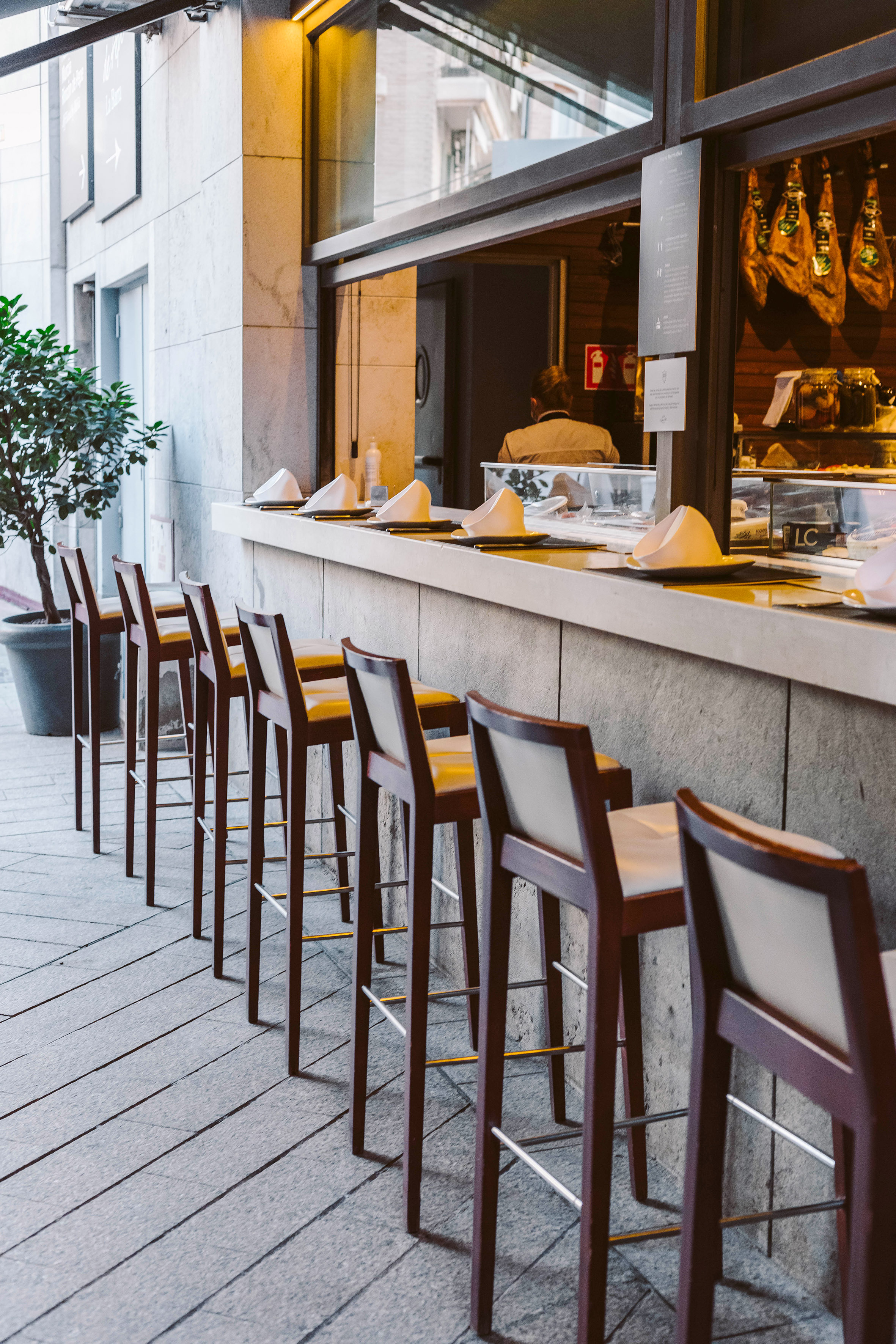 Outdoor bar stools at a street cafe counter with hanging hams inside. - La Barra del Rincón Restaurant, Tapas - Hotel Murcia Rincón de Pepe Affiliated by Meliá, Murcia