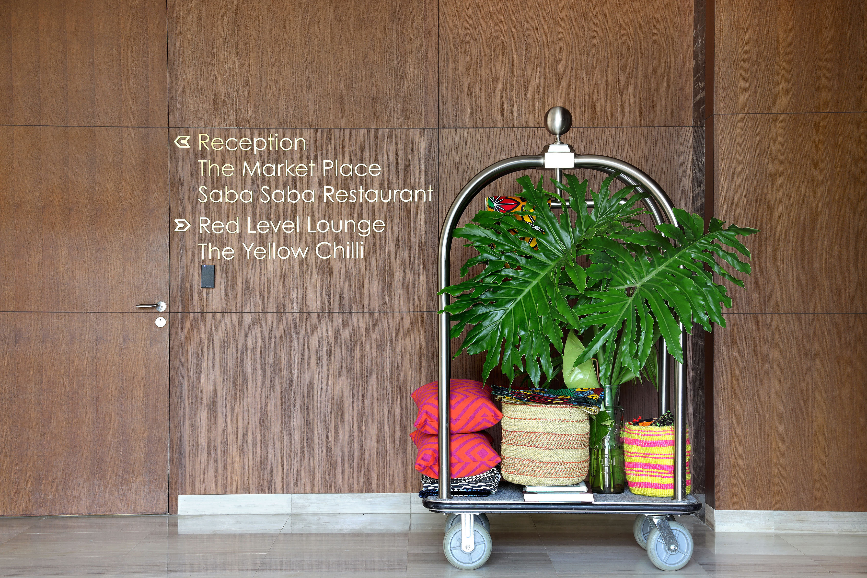 Hotel lobby sign to reception, restaurants, lounge; bell cart with plant decor.