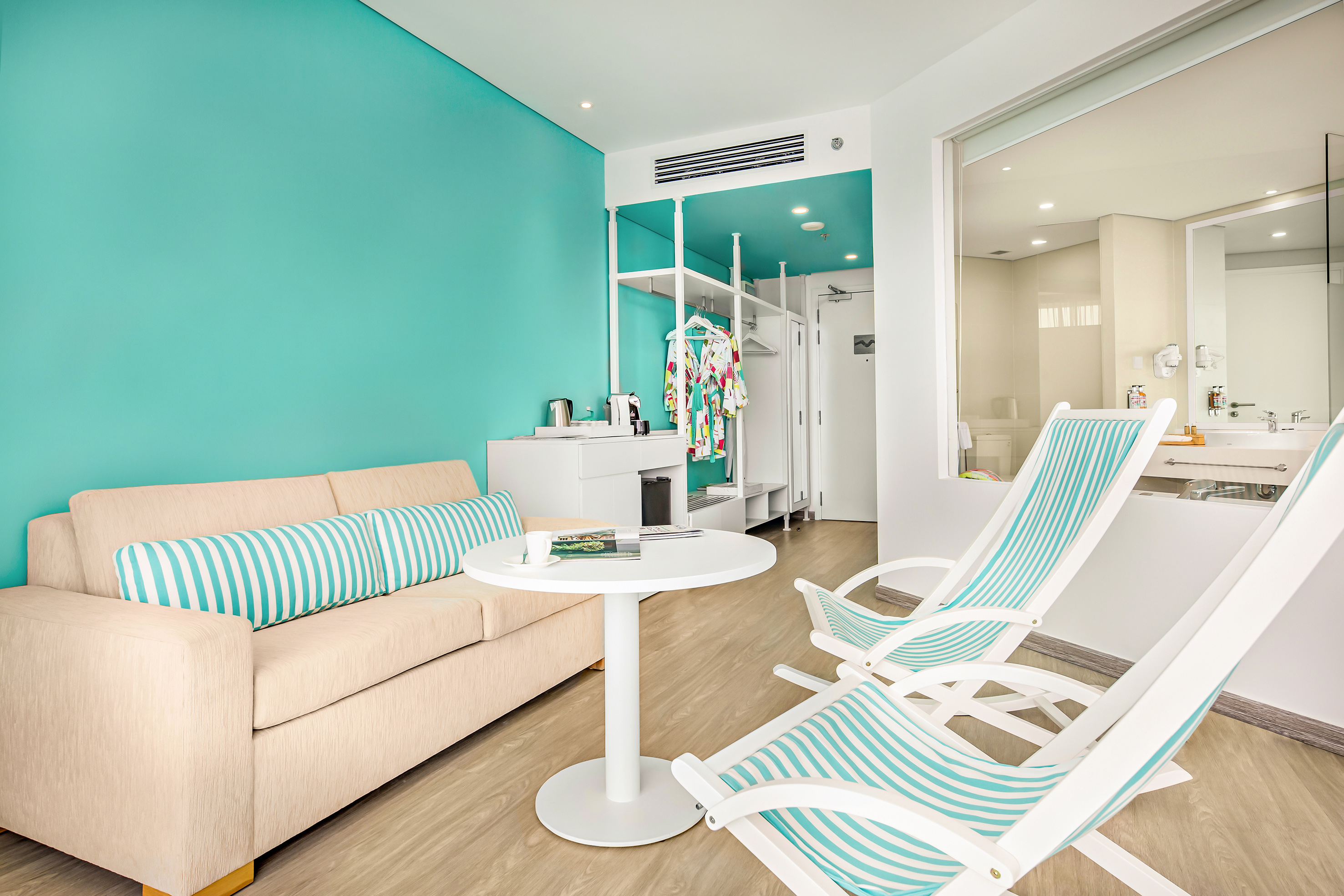 Tropical hotel room with teal accent wall, beige sofa, and striped lounge chairs.