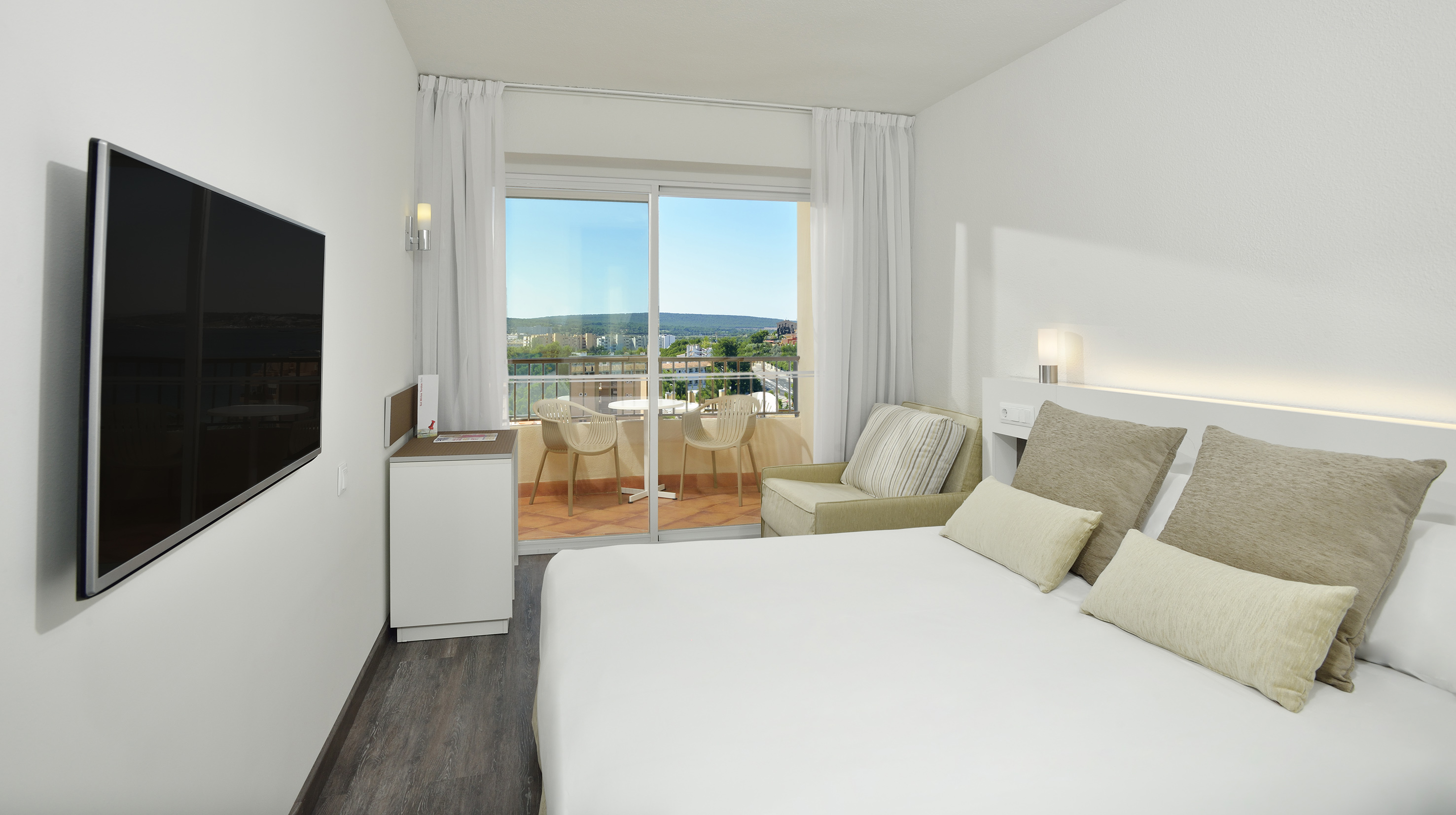 Modern hotel room with bed, TV, and balcony overlooking a city landscape.
