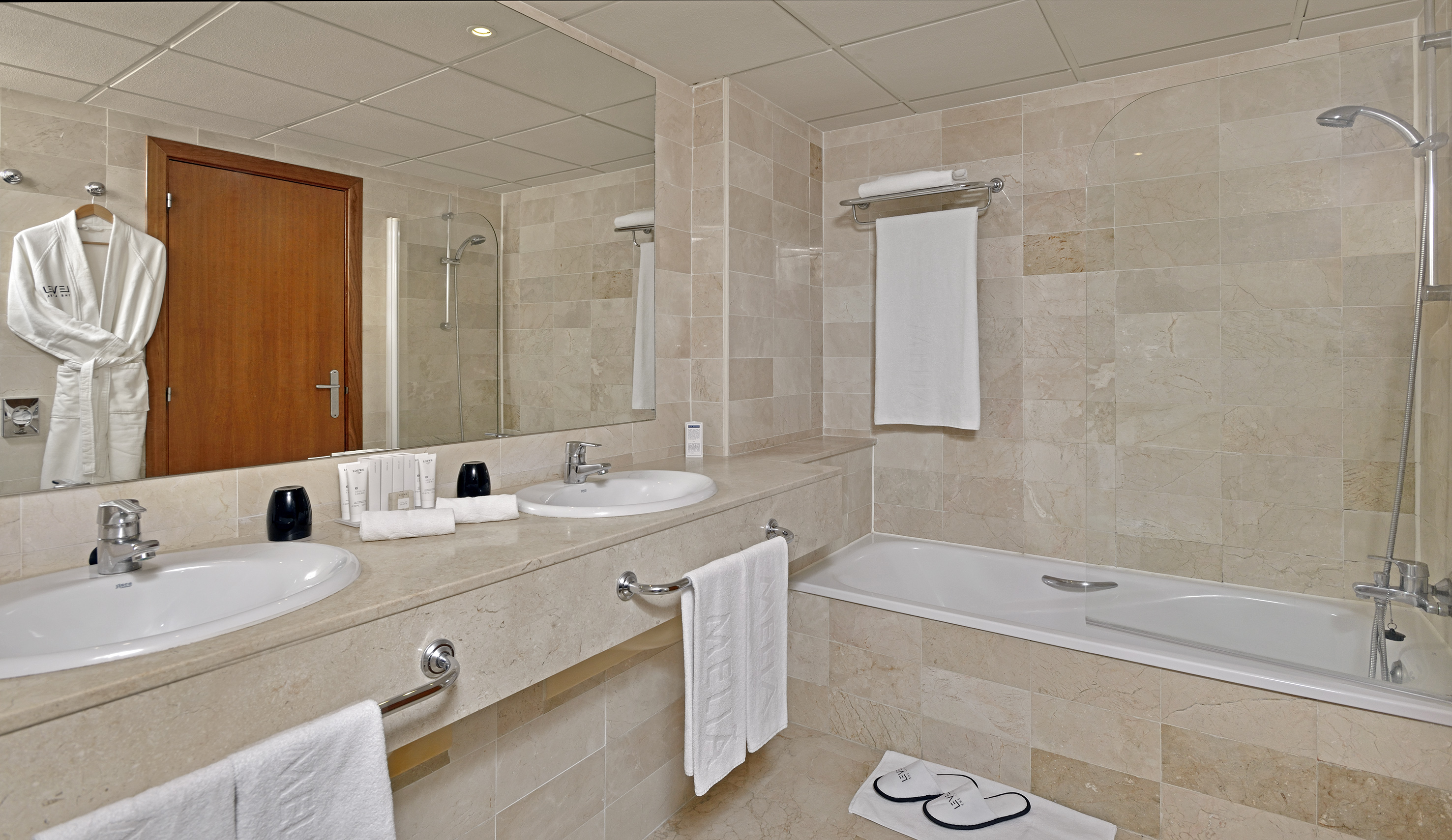 Spacious hotel bathroom with dual sinks, large mirror, bathtub, and marble tiles.