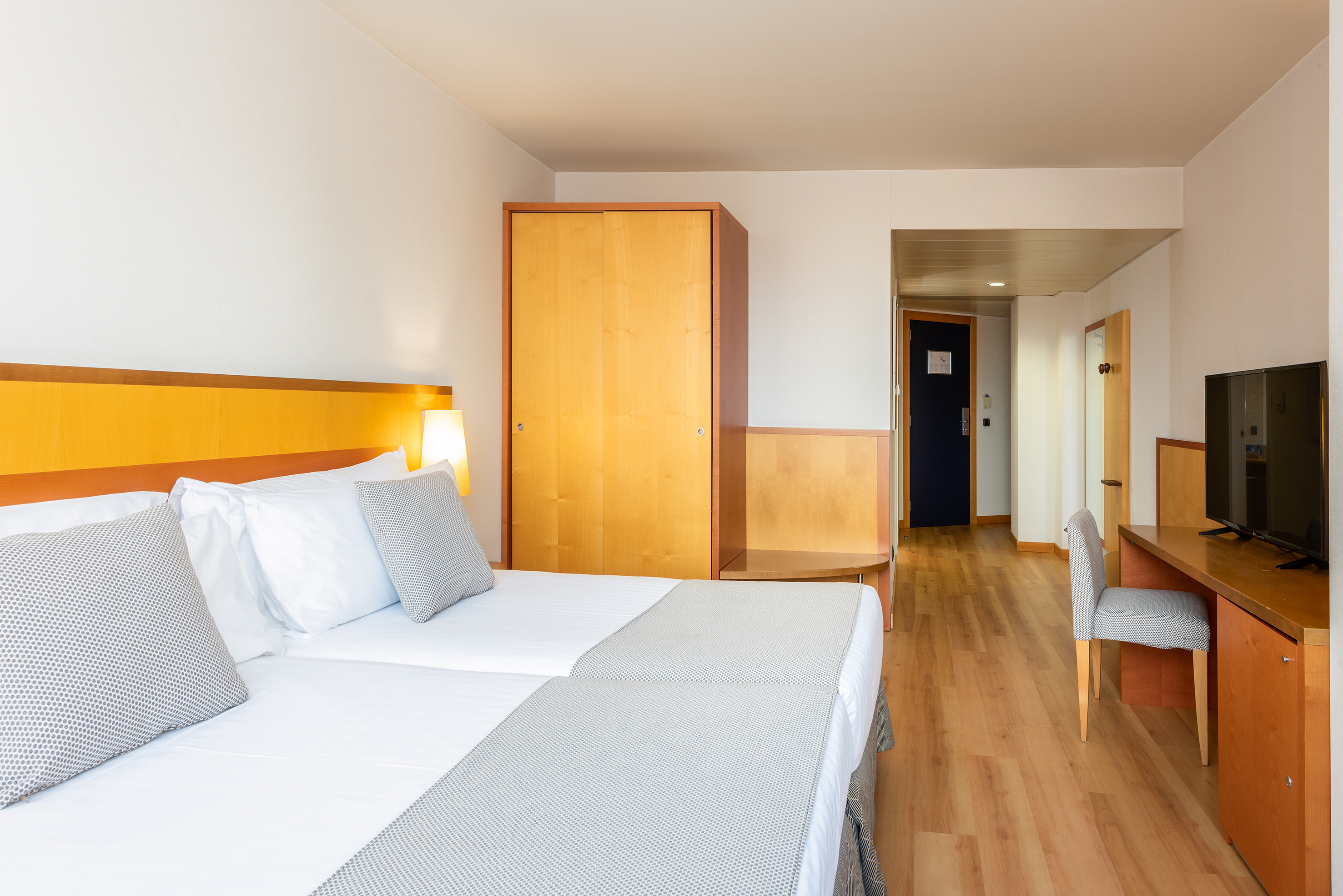 Bright hotel room with twin beds, wooden wardrobe, and desk with TV.