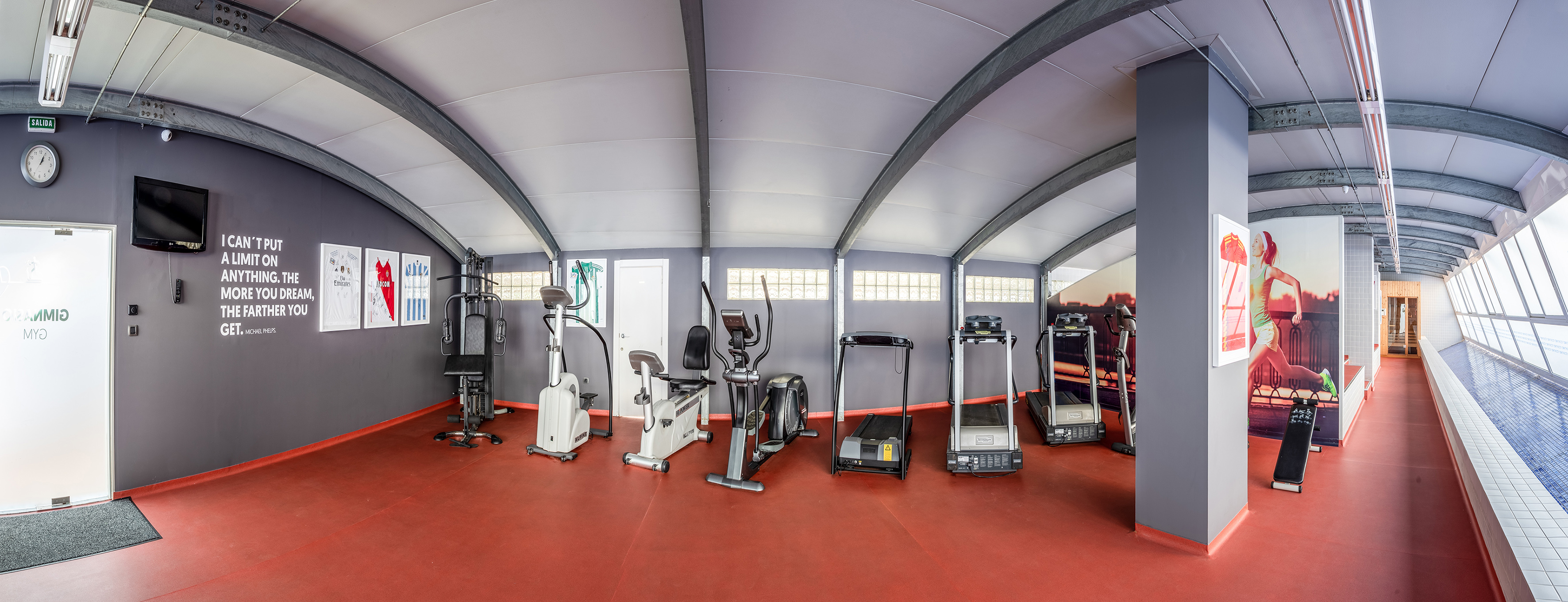 Spacious gym with various exercise machines, red floor, and an indoor pool.
