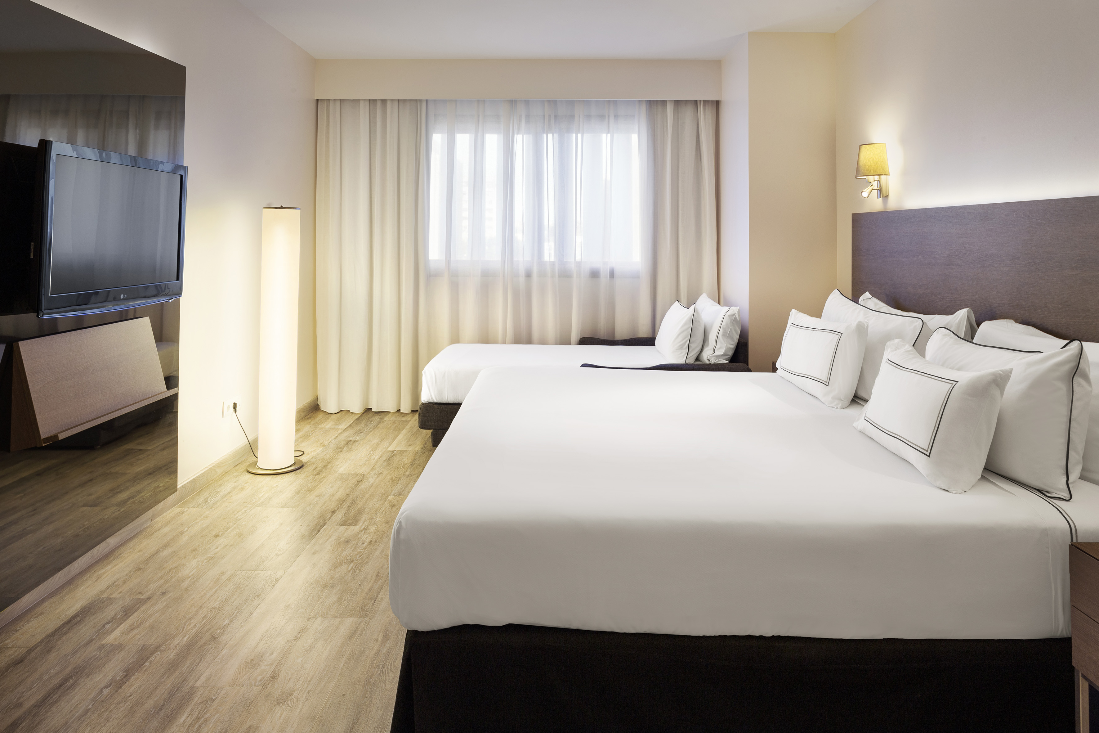 Modern hotel room with king bed, sofa bed, TV, and light wood floor.