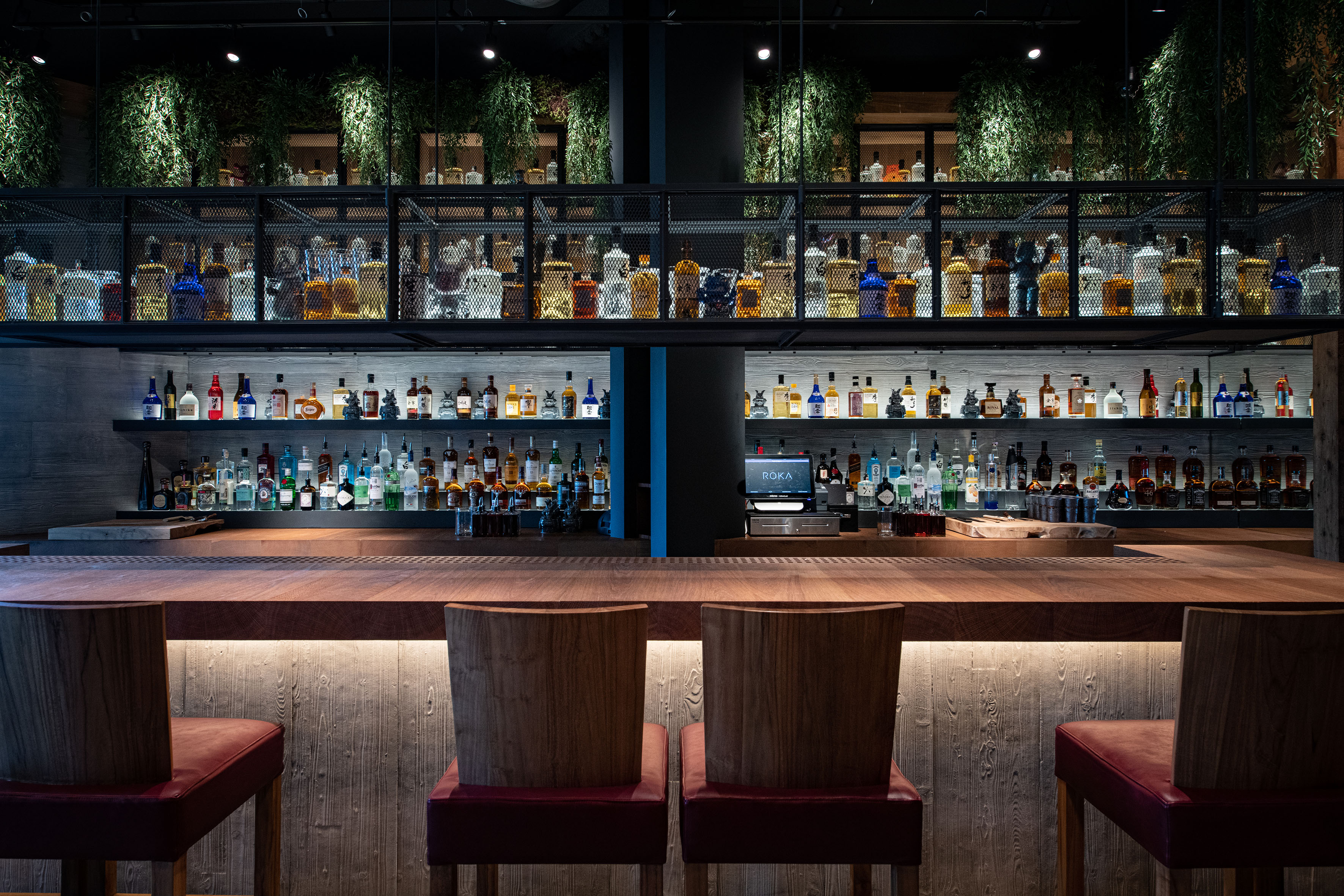 ROKA bar with glowing liquor bottles, dark wood counter, and red stools.