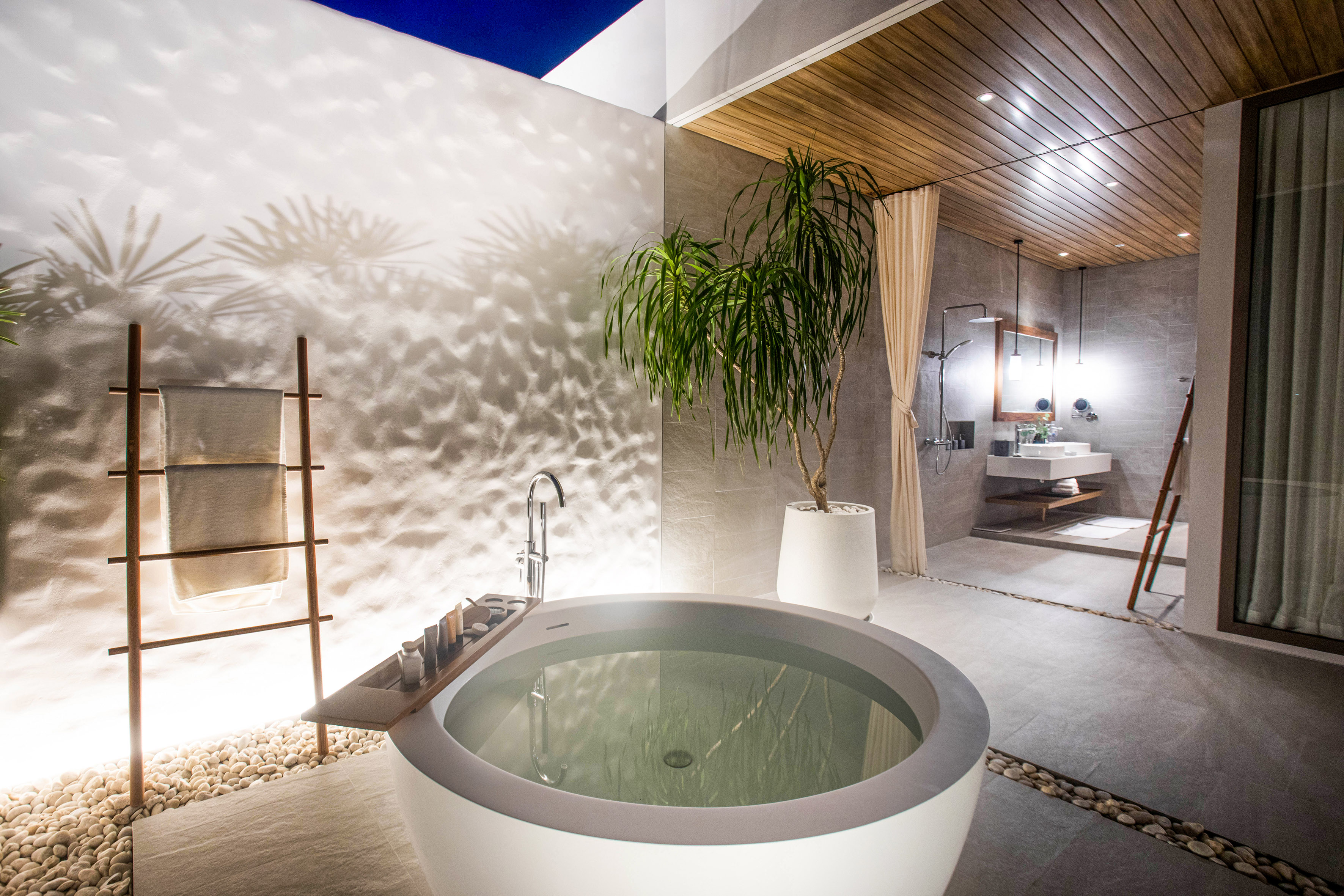 Modern outdoor bathroom with round freestanding tub, pebble floor, and plant.