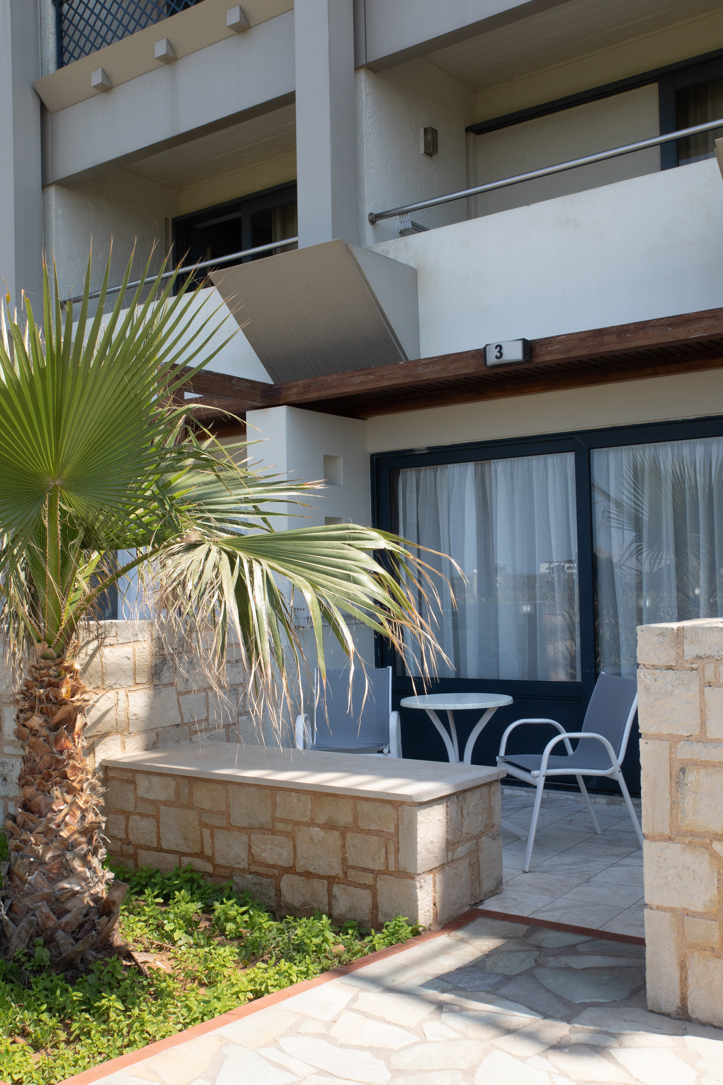 Sunny hotel terrace with palm tree, stone seating, table, and chairs.