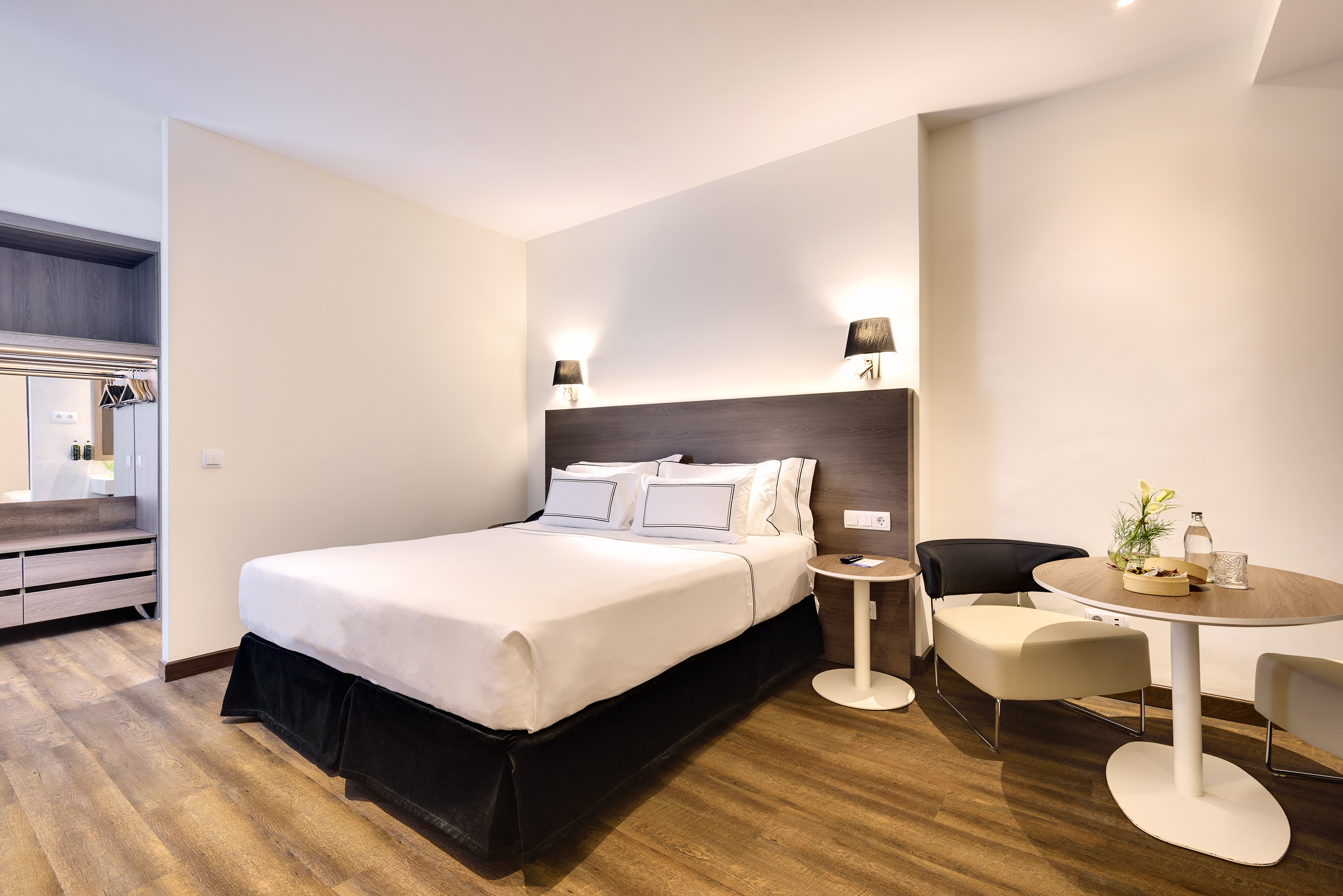 Modern hotel bedroom with queen bed, dark headboard, wood floor, and seating area.